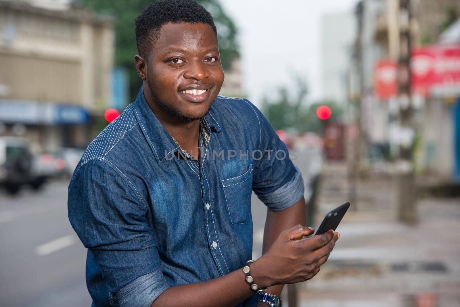 smiling young man sitting outside on an iron bar and holding his phone in hands at the end of the day