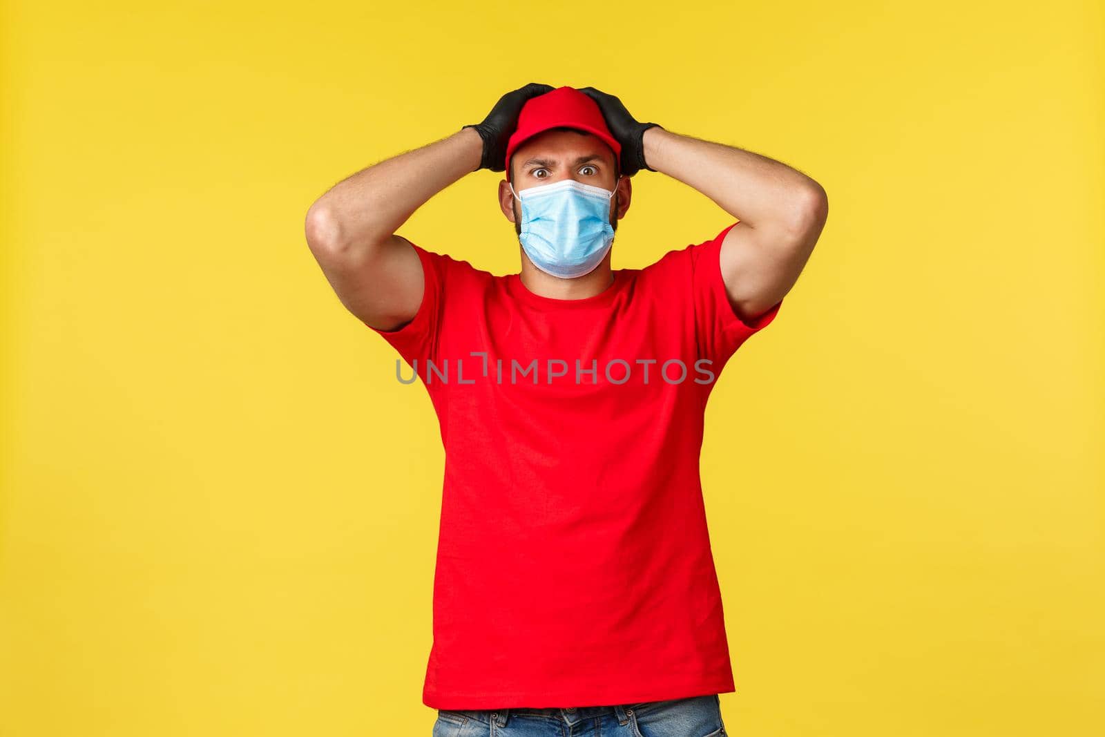 Express delivery during pandemic, covid-19, safe shipping, online shopping concept. Nervous and concerned courier in red uniform, medical mask, staring shocked, feel trouble, grab head.