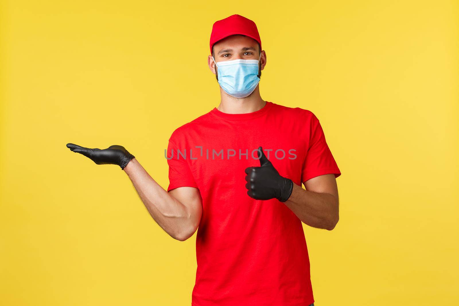 Express delivery during pandemic, covid-19, safe shipping, online shopping concept. Smiling courier in red uniform, medical mask and gloves, showing product and thumb-up in approval.