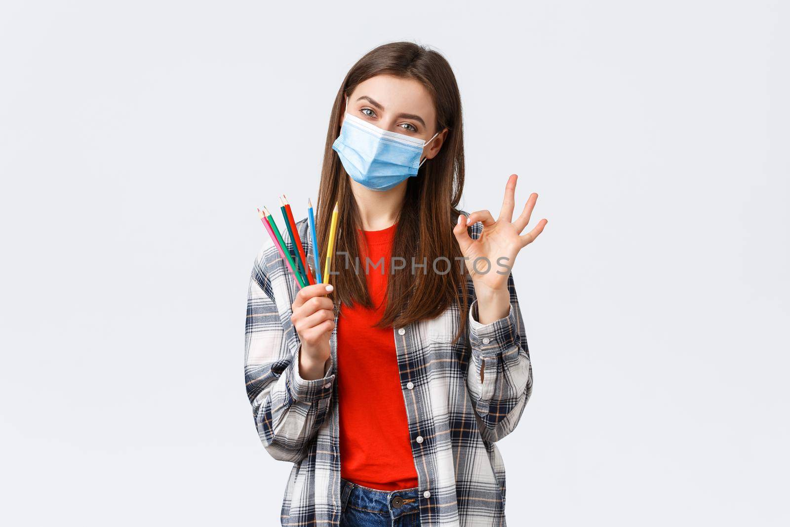 Social distancing, hobbies on covid-19 outbreak, coronavirus concept. Cute cheerful girl in medical mask make online lesson on how to draw during self-quarantine, show colored pencils and OK sign.