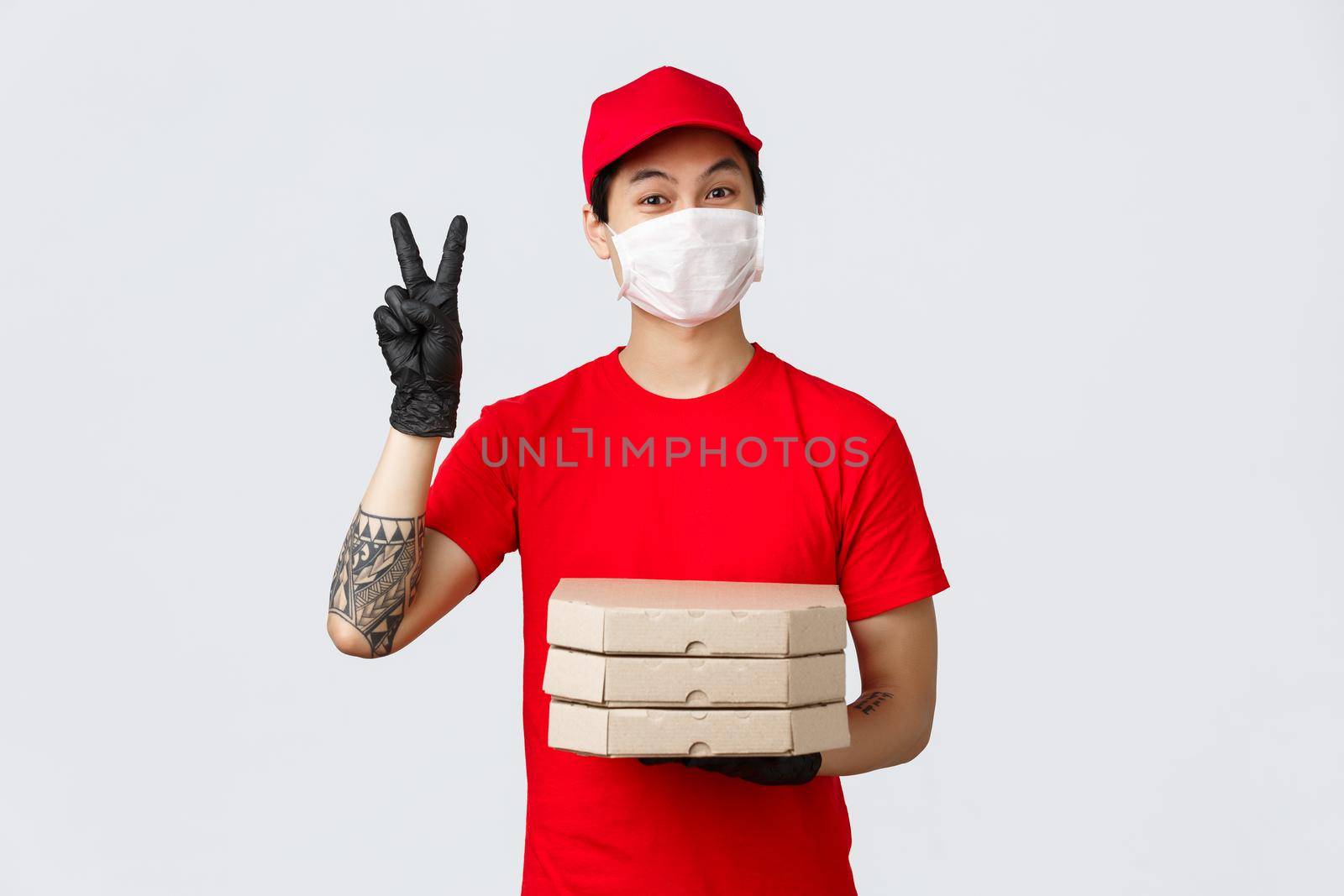 Cute asian delivery man in medical mask and gloves, wearing red uniform cap, holding pizza boxes and make peace sign, see client, suggest contactless deliver food during virus outbreak, covid 19.