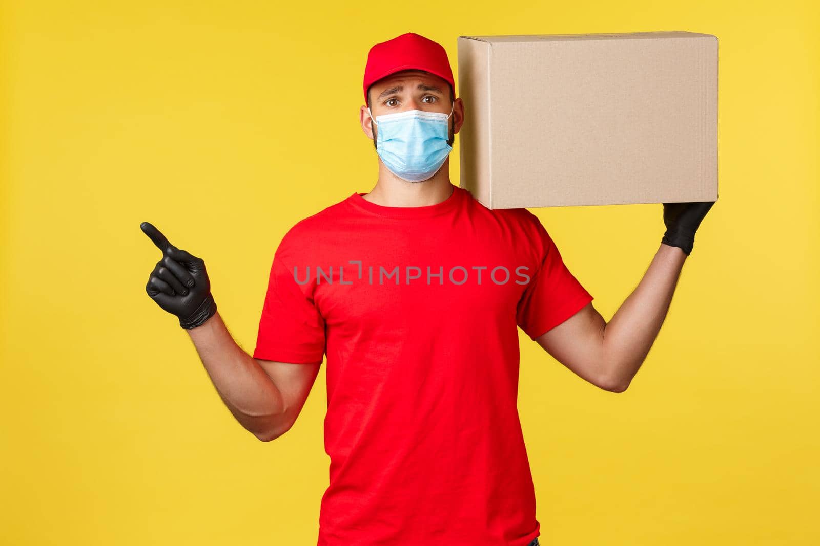 Express delivery during pandemic, covid-19, safe shipping, online shopping concept. Upset and sad courier in red uniform and medical mask, pointing finger left, hold box package on shoulder.