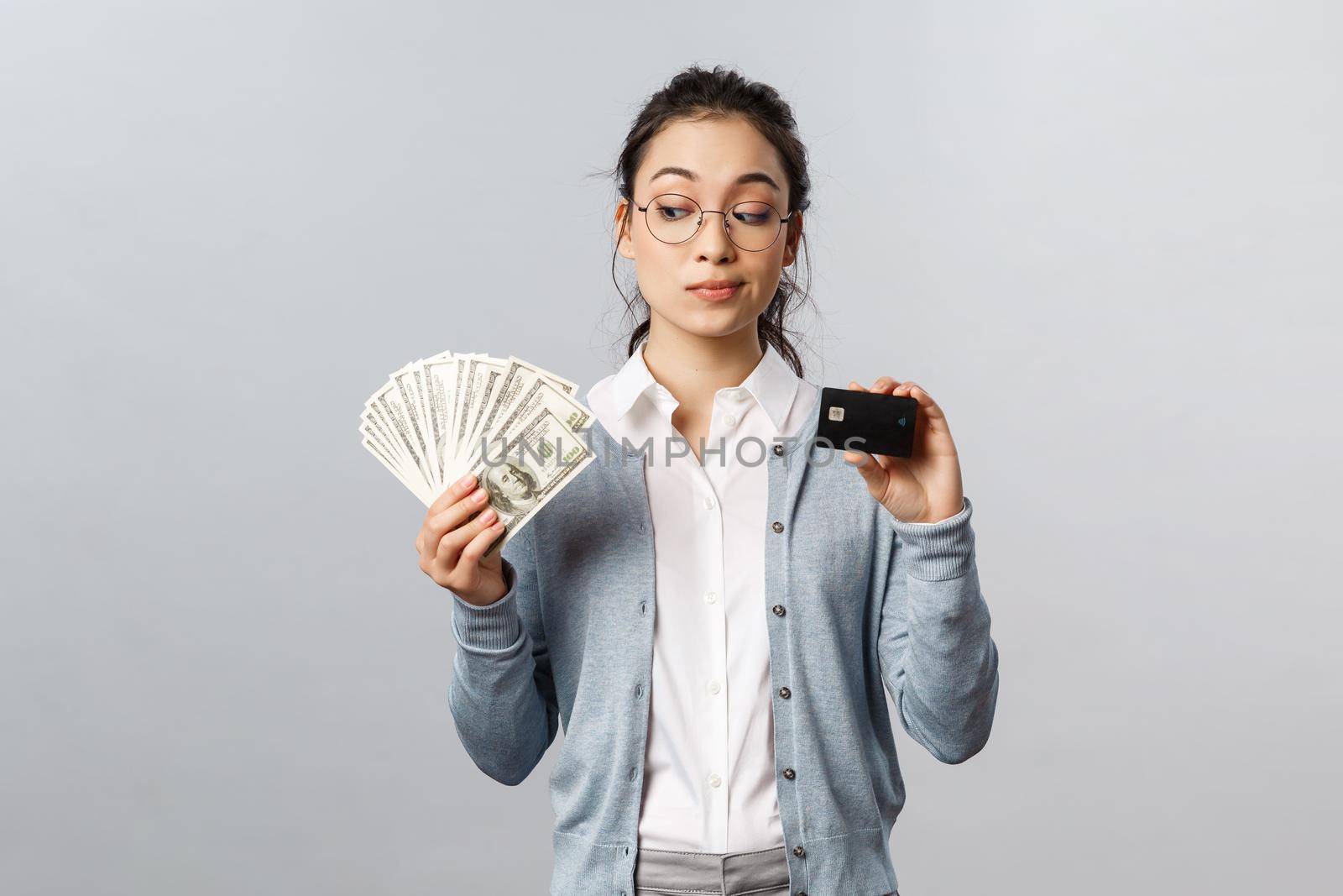 Business, finance and investment concept. Thoughtful indecisive cute asian girl look tempting at cash dollars, holding money and credit card, shopping, think where invest, grey background.