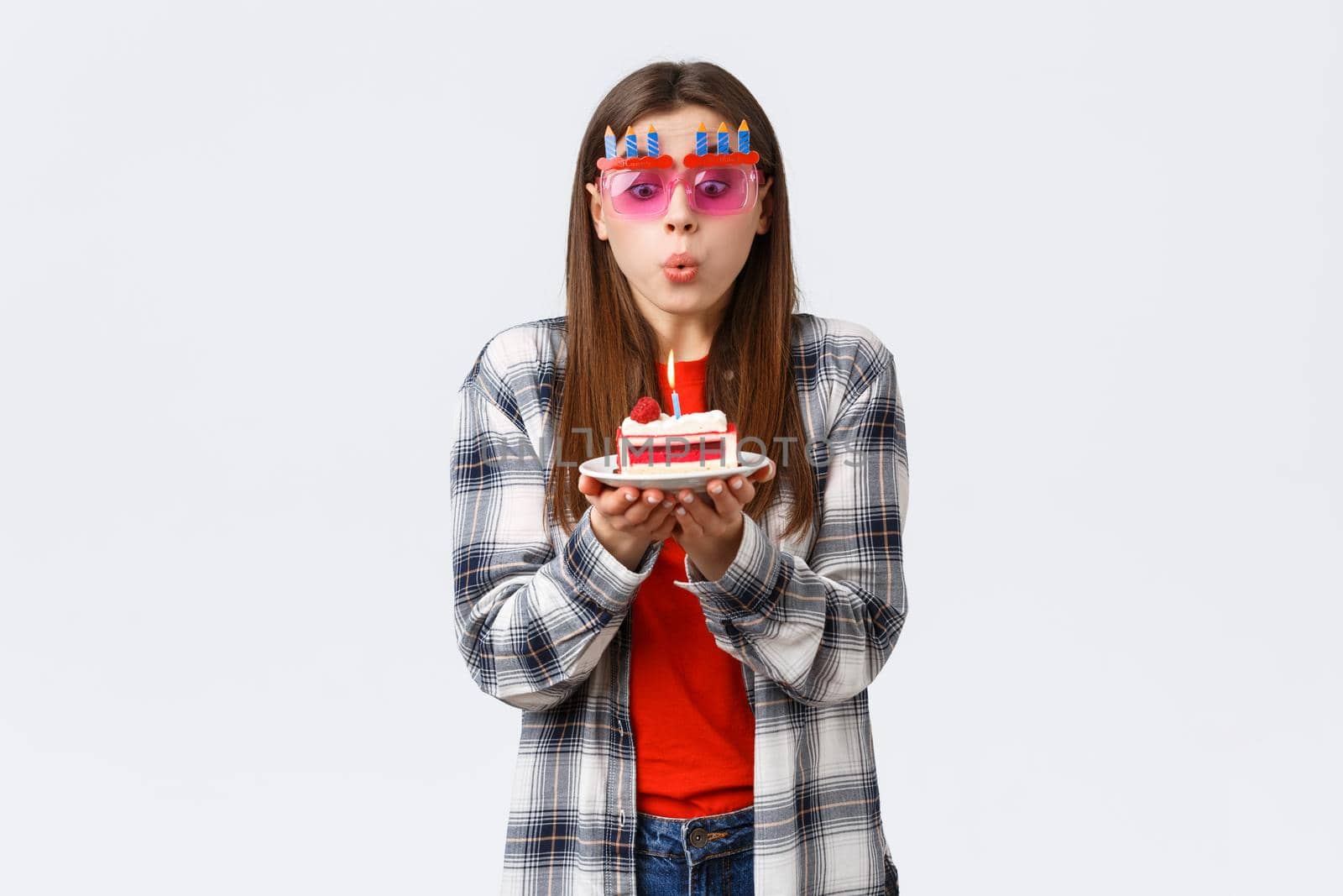 People lifestyle, holidays and celebration, emotions concept. Cute tender young b-day girl in glasses, blowing lit candle on birthday cake, making wish and celebrating at party, white background by Benzoix