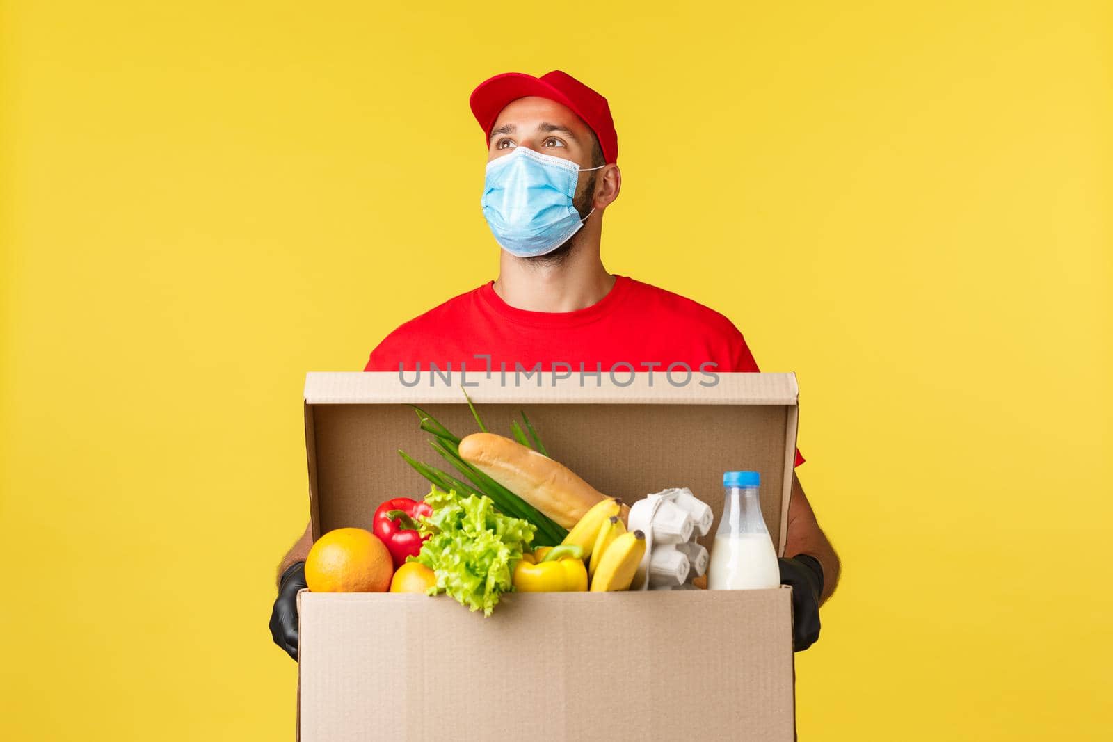 Takeaway delivery, covid-19 quarantine and groceries concept. Young courier in red cap and t-shirt uniform, medical mask, look up thoughtful, searching customer apartment, hold box with food order.