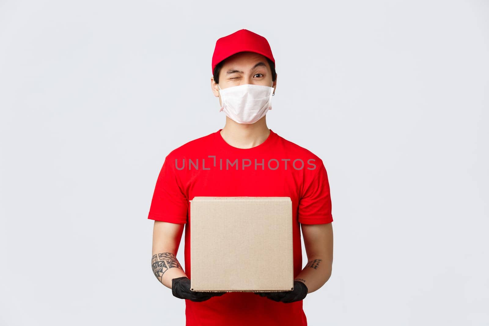 Friendly asian delivery man in red cap and t-shirt, wear protective gloves and medical mask for client and employees safety, house delivery during covid-19 quarantine, holdin box package.