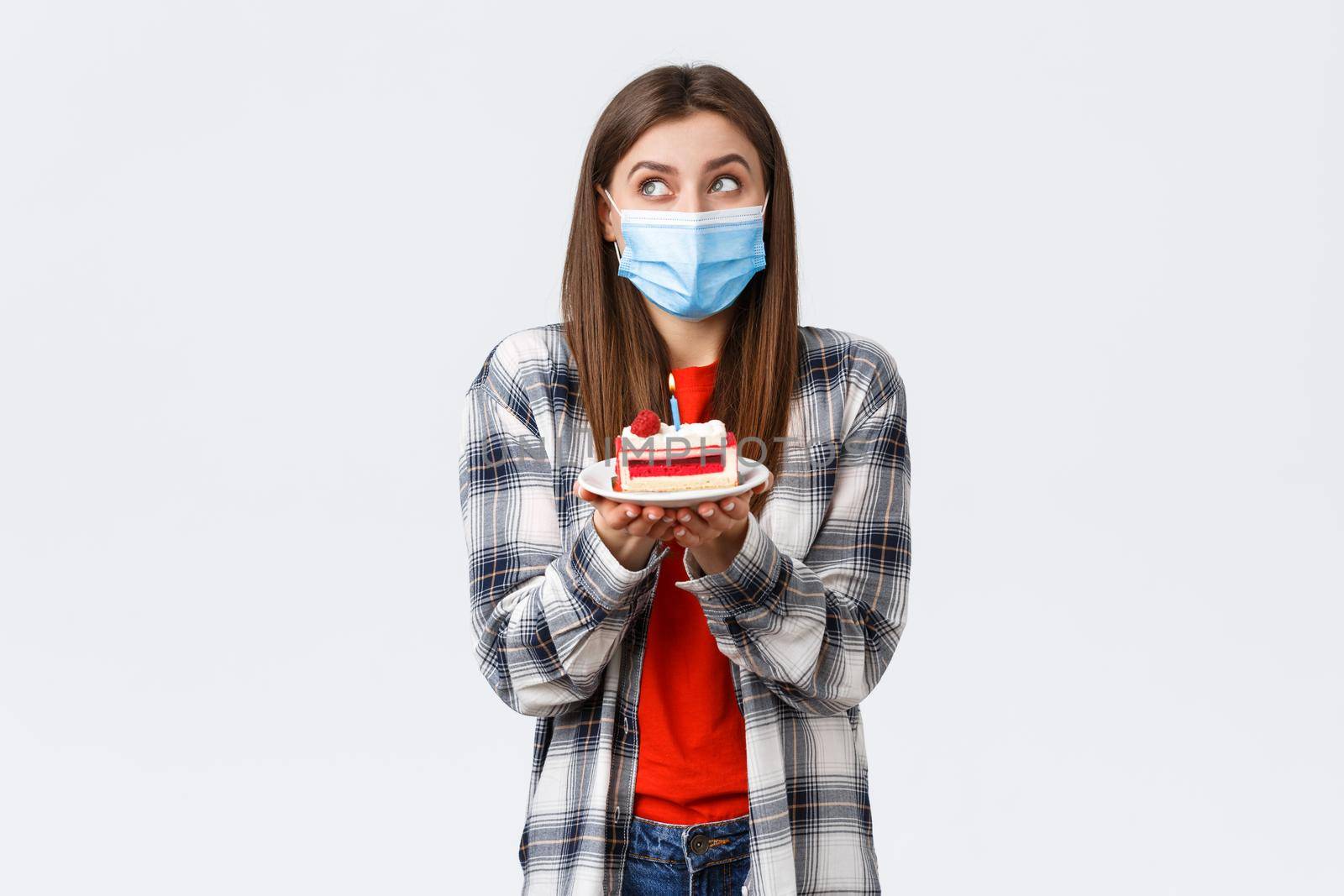 Coronavirus outbreak, lifestyle during social distancing and holidays celebration concept. Dreamy cute girl in medical mask, dreamy look up, imaging making wish as holding birthday cake with candle by Benzoix