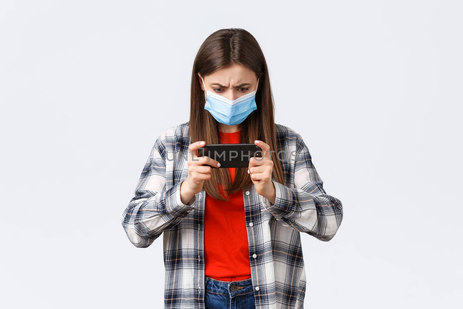 Different emotions, covid-19 pandemic, coronavirus self-quarantine and social distancing concept. Serious focused young girl in medical mask trying pass level in mobile game, tap screen and frowning.