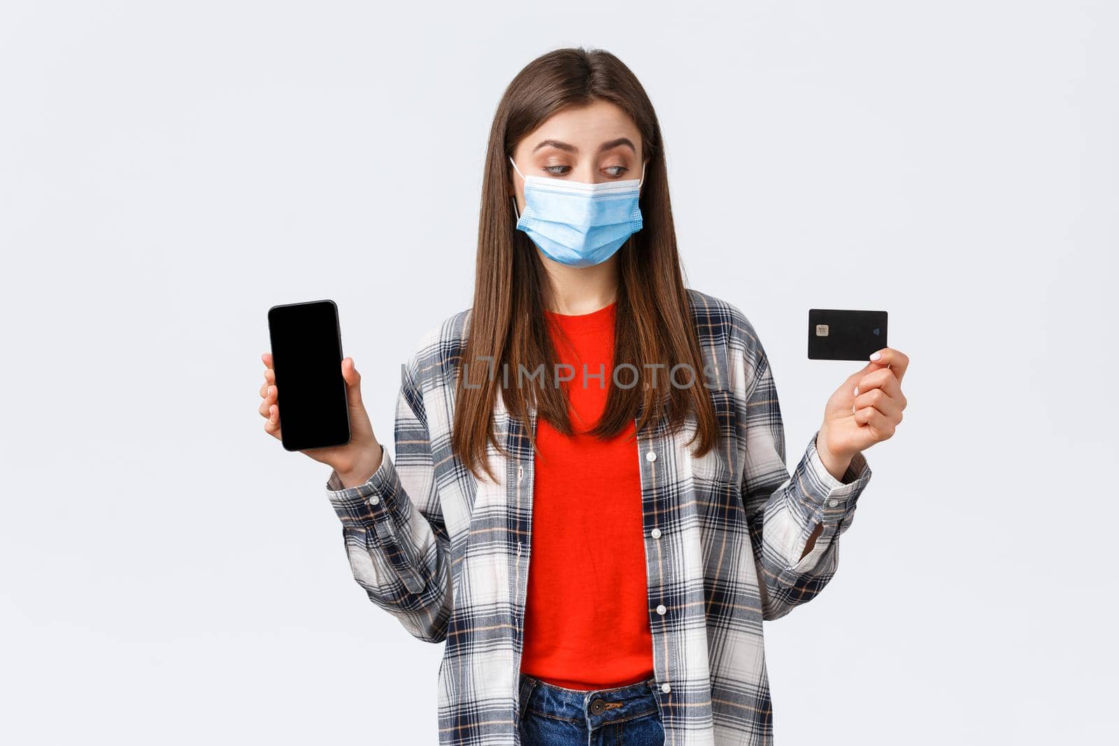 Coronavirus outbreak, working from home, online shopping and contactless payment concept. Girl in medical mask look at credit card as show mobile phone screen, making order by Benzoix