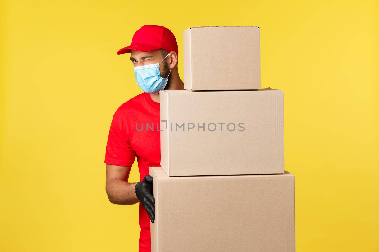 Express delivery during pandemic, covid-19, safe shipping, shopping concept. Friendly handsome courier in red uniform, medical mask, standing near pile of box orders, taking care customers packages.