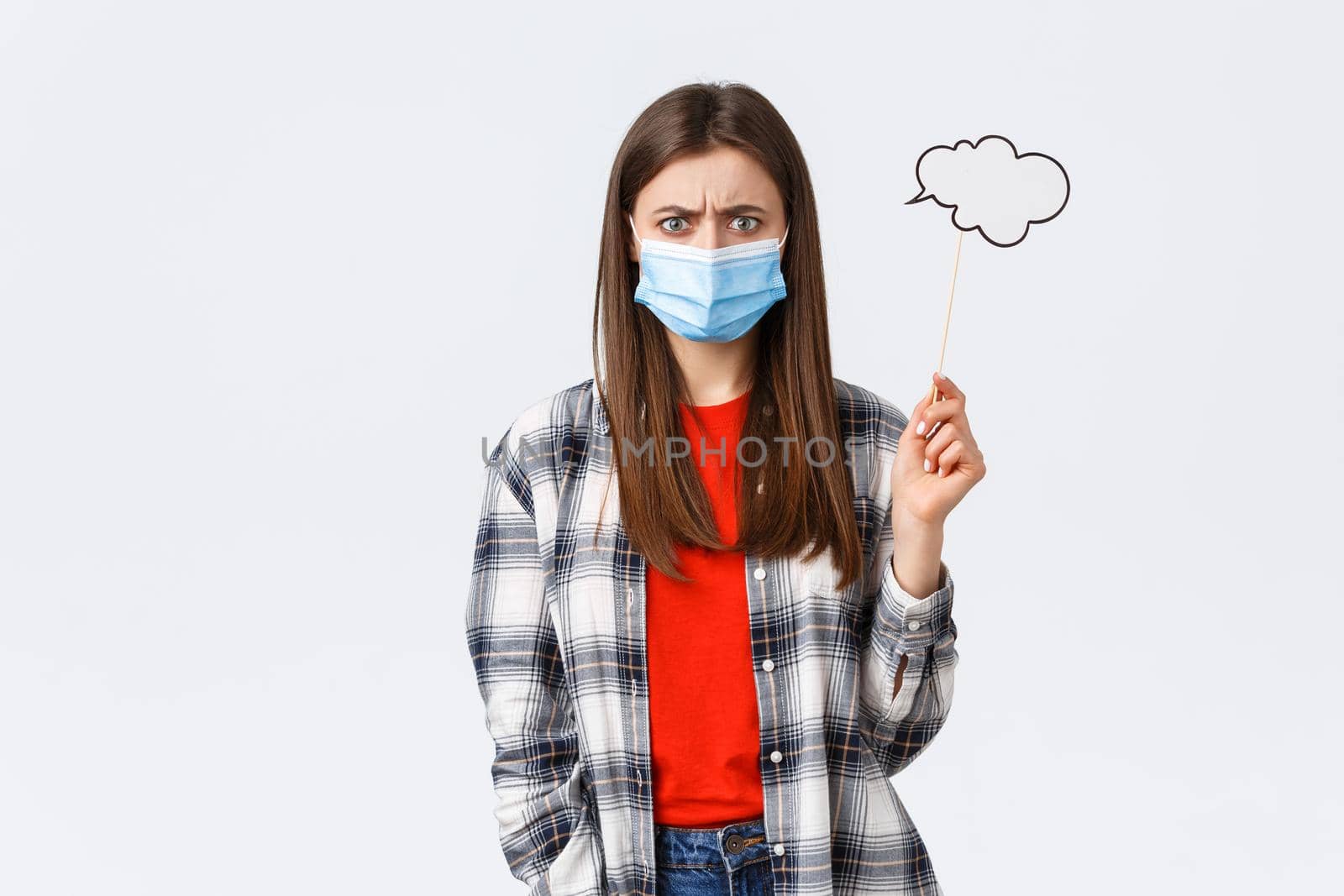 Coronavirus outbreak, leisure on quarantine, social distancing and emotions concept. Troubled young woman in medical mask frowing upset or disappointed, hold comment cloud stick near head.