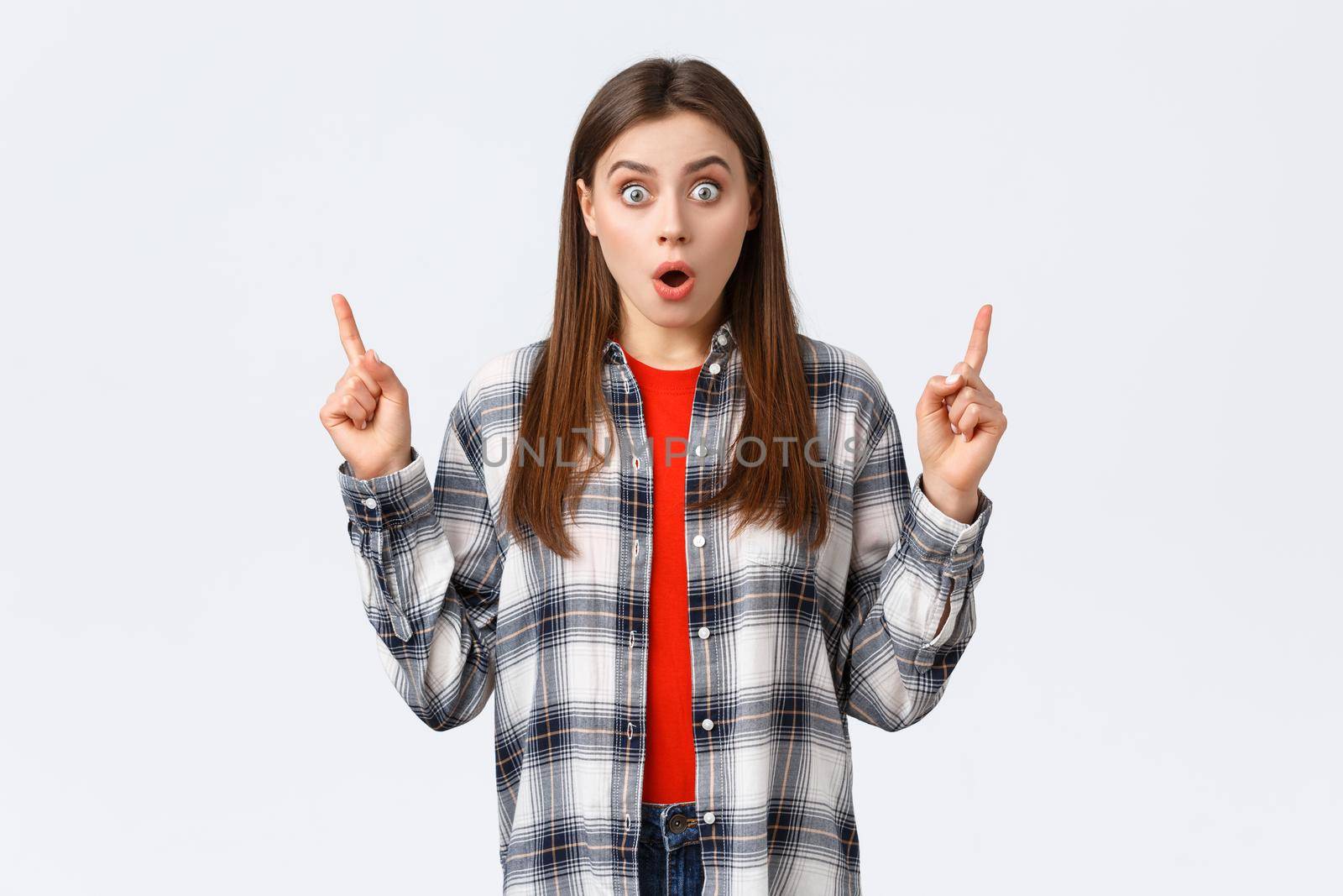 Lifestyle, different emotions, leisure activities concept. Astonished and speechless attractive girl in checked shirt pointing fingers up to tell news, drop jaw staring, make wow expression by Benzoix