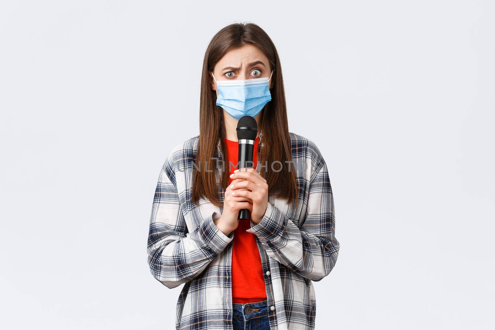 Coronavirus outbreak, leisure on quarantine, social distancing and emotions concept. Confused woman in medical mask look smth strange, holding microphone, perform, white background.