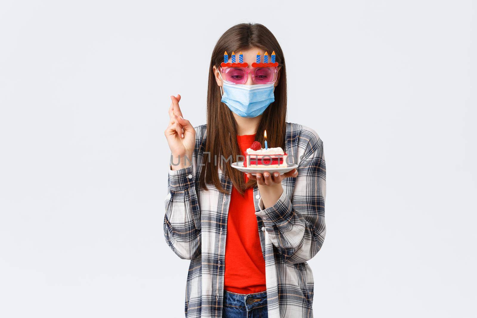 Coronavirus outbreak, lifestyle social distancing and celebration concept. Enthusiastic happy girl in glasses and medical mask celebrate birthday on quarantine, cross fingers, make wish at cake by Benzoix