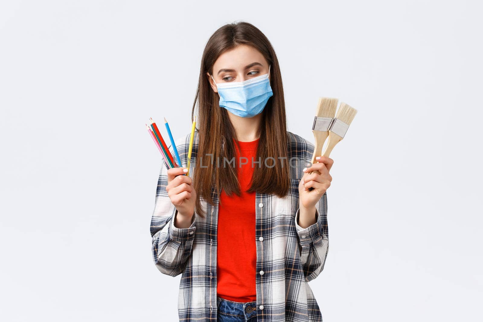 Social distancing, leisure and hobbies on covid-19 outbreak, coronavirus concept. Pleased smiling girl in medical mask start draw, look at colored pencils, show painting brushes.