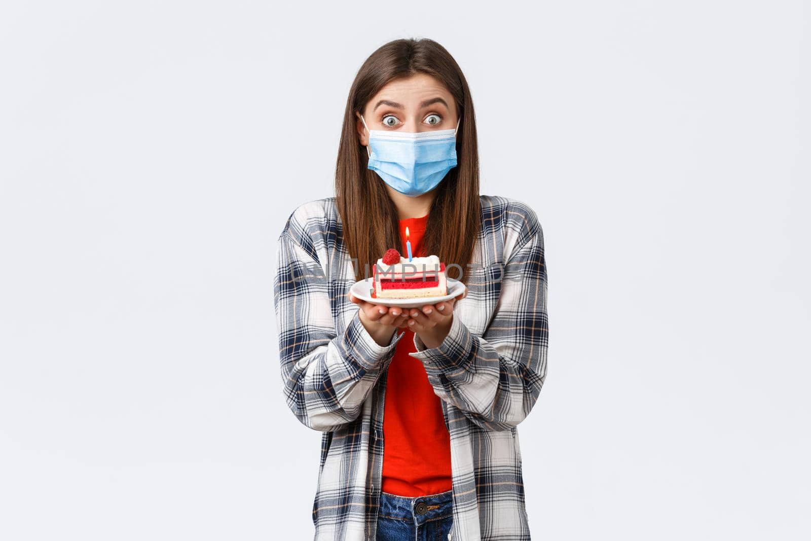 Coronavirus outbreak, lifestyle during social distancing and holidays celebration concept. Cute happy birthday girl making wish, wear medical mask, holding b-day cake, celebrating inside home.