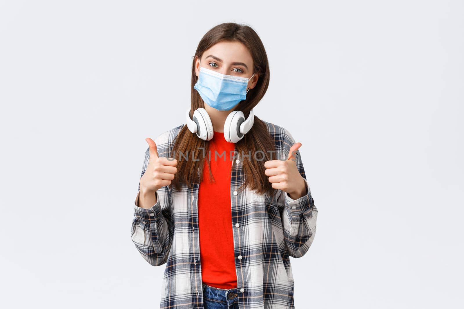 Social distancing, leisure and lifestyle on covid-19 outbreak, coronavirus concept. Cute pleased young caucasian woman in medical mask, take-off headphones to say yes or good, make thumb-up sign by Benzoix