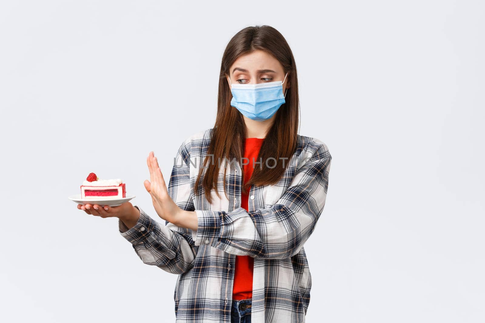 Coronavirus outbreak, lifestyle during social distancing and holidays celebration concept. Displeased and reluctant cute girl in medical mask refuse eat cake, show stop sign at dessert by Benzoix