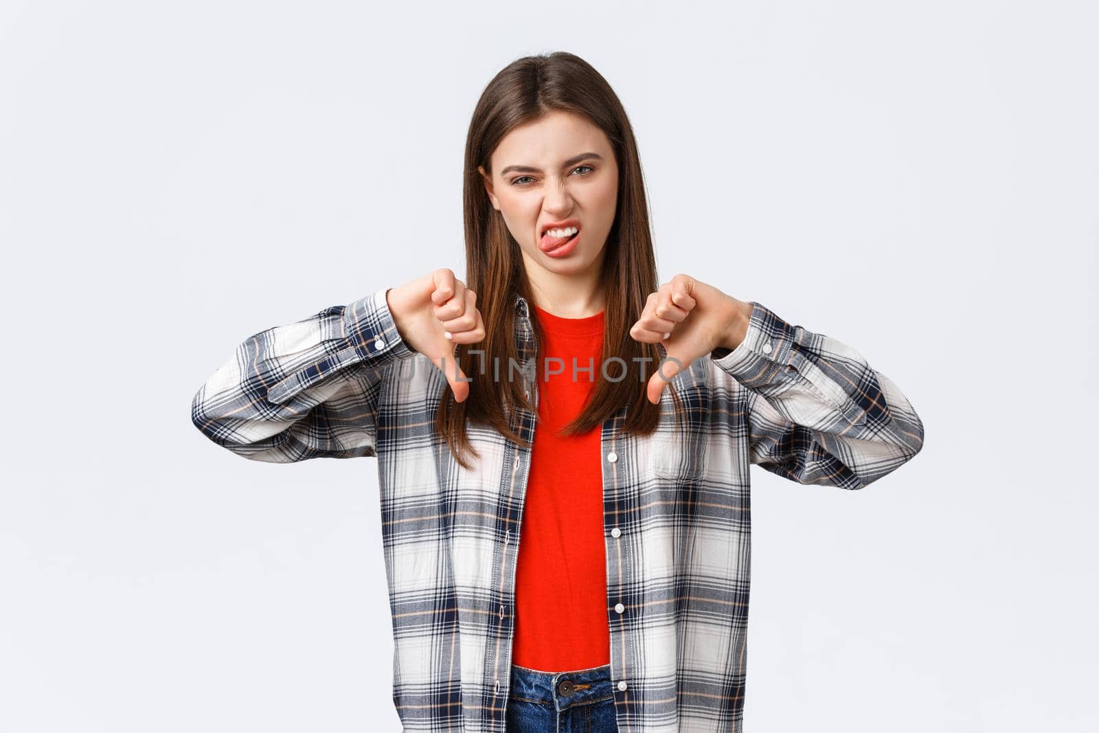 Lifestyle, different emotions, leisure activities concept. Displeased and unimpressed young picky girl in checked shirt, thumb-down and show tongue to express dislike, rate awful movie.