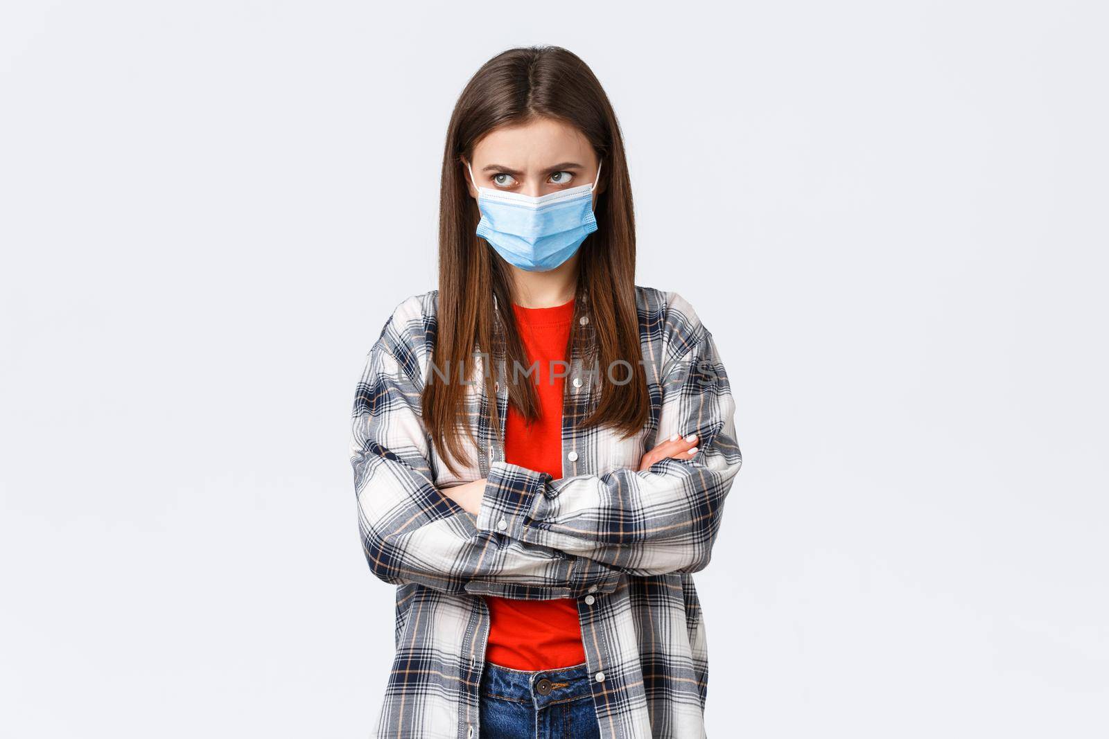 Coronavirus outbreak, leisure quarantine, social distancing and emotions concept. Mad young cute girl in medical mask, cross arms chest, look from under forehead upper left corner, jealous or angry.