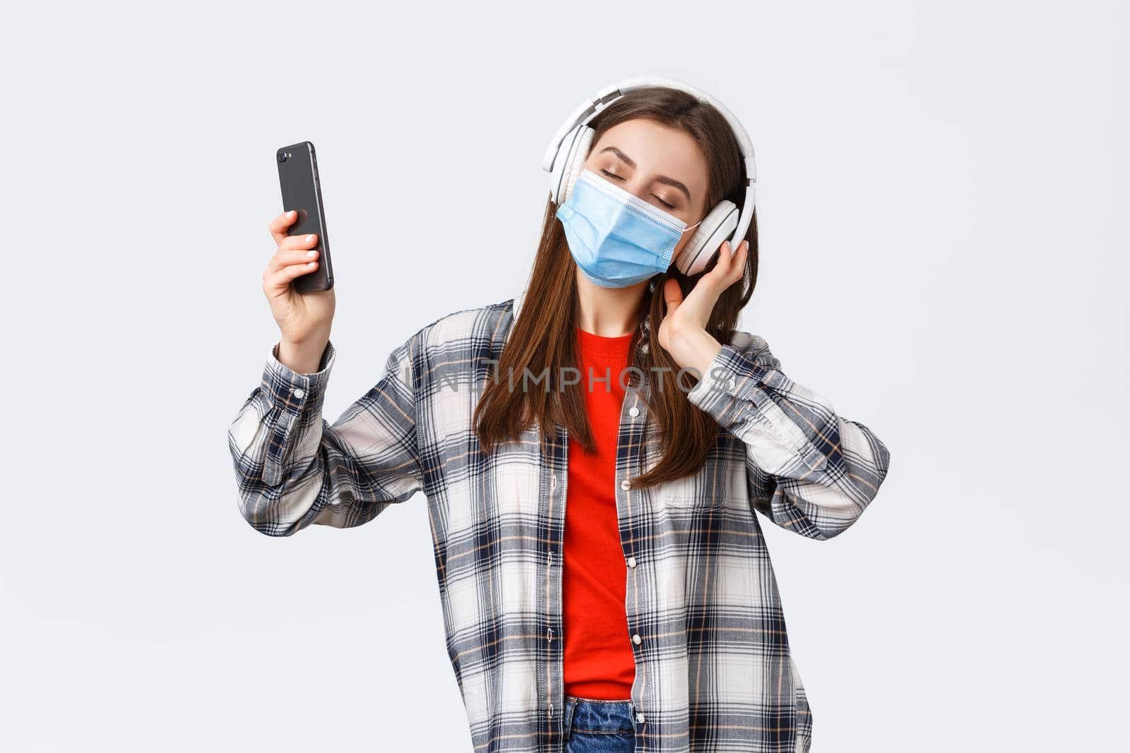 Social distancing, leisure and lifestyle on covid-19 outbreak, coronavirus concept. Carefree young girl in medical mask and headphones, close eyes dancing to music, perfect sound, hold mobile phone.