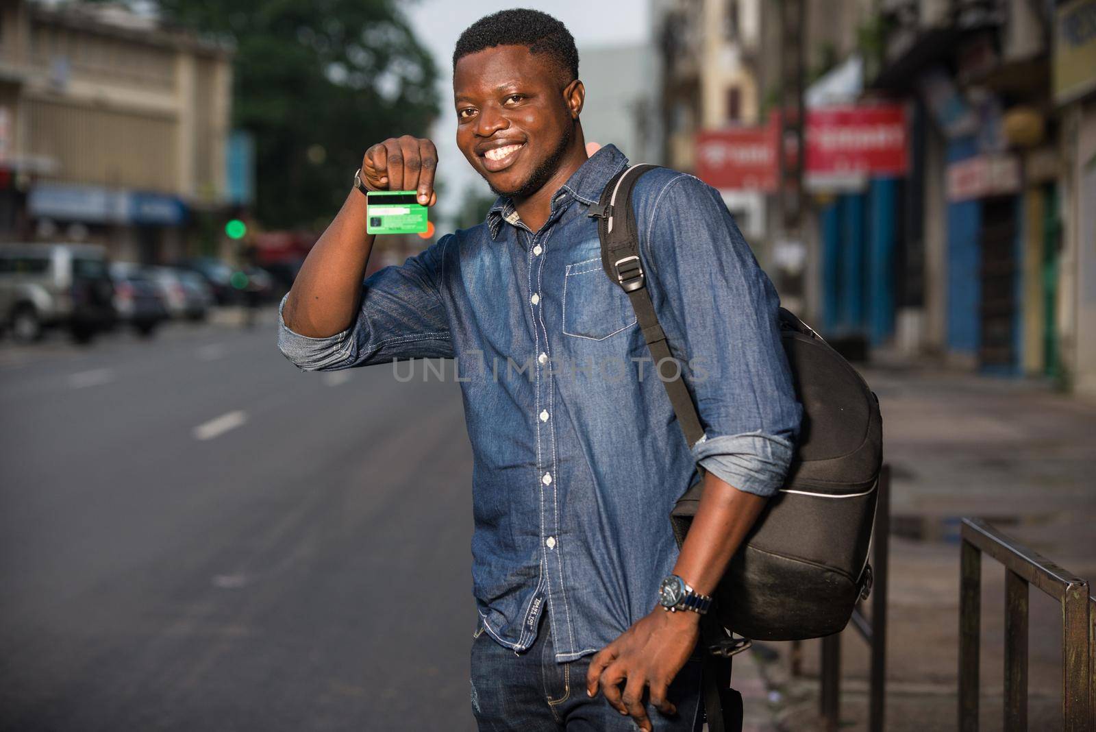 Young single traveler on the street at the platform of the station with backpack. Smiling man showing credit card, waiting for vehicle at train station for trip. Summer vacation traveling or young tourist concept