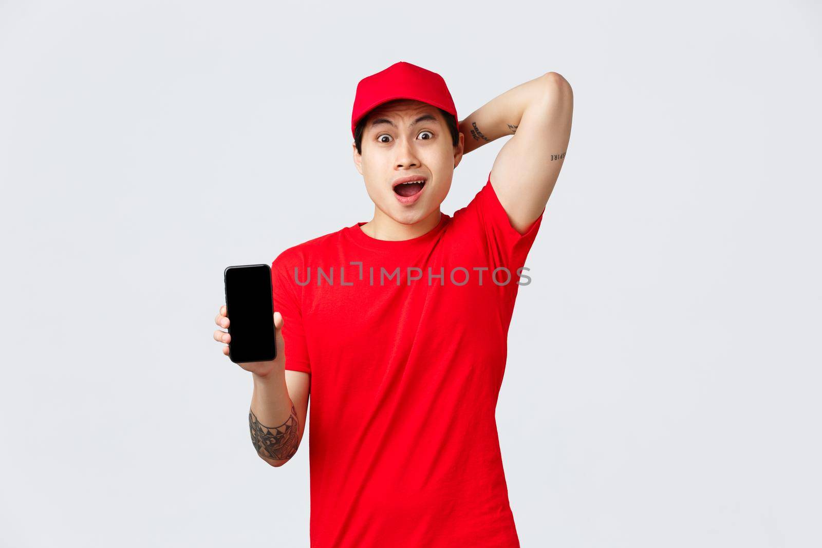 Nervous and troubled asian delivery guy in red cap and t-shirt uniform, gasping anxious, look confused and puzzled showing smartphone screen bad news, mixed-up order, stand grey background.