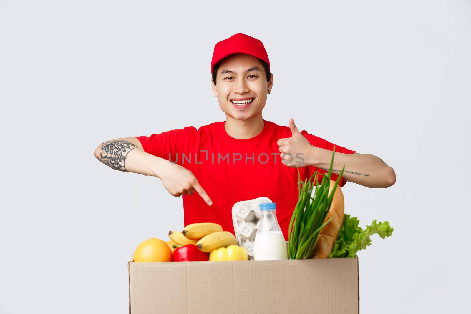 Online shopping, food delivery and internet stores concept. Smiling friendly courier in red t-shirt and cap, show thumb-up, point at order grocery package, point products, recommend carrier service.