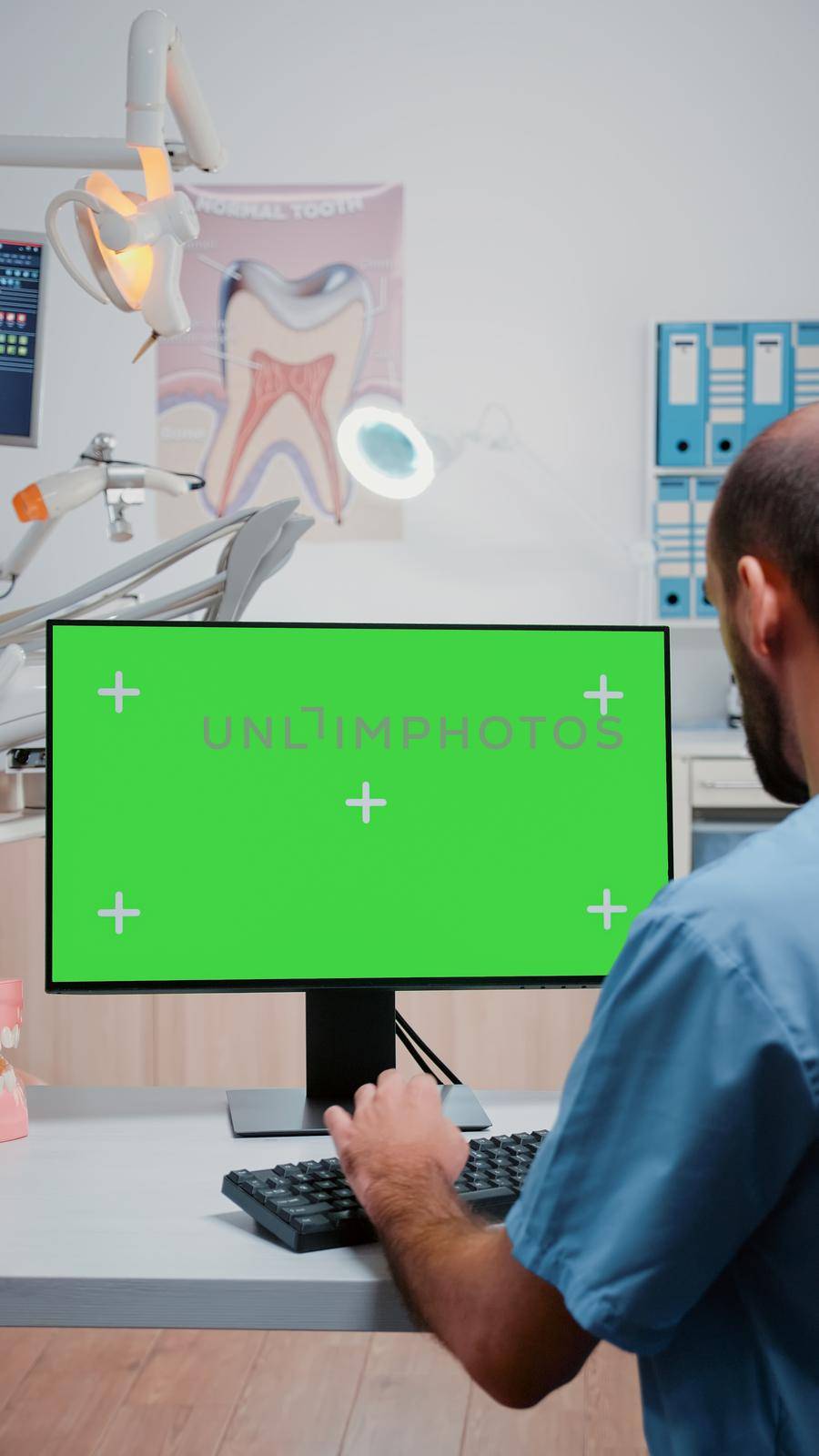 Vertical video: Assistant looking at horizontal green screen on computer at dentist office. Man working as teethcare specialist with mockup background and isolated template on monitor