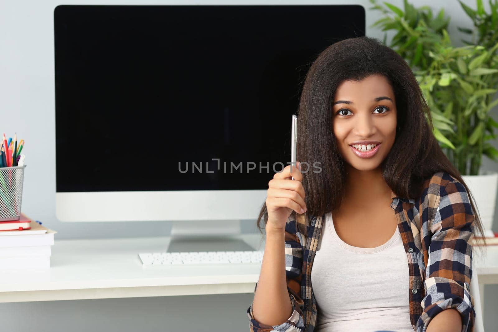 Portrait of afro american young lady sit in office with computer device behind on desk. Cute woman with curly hairstyle hold silver pen. Secretary concept