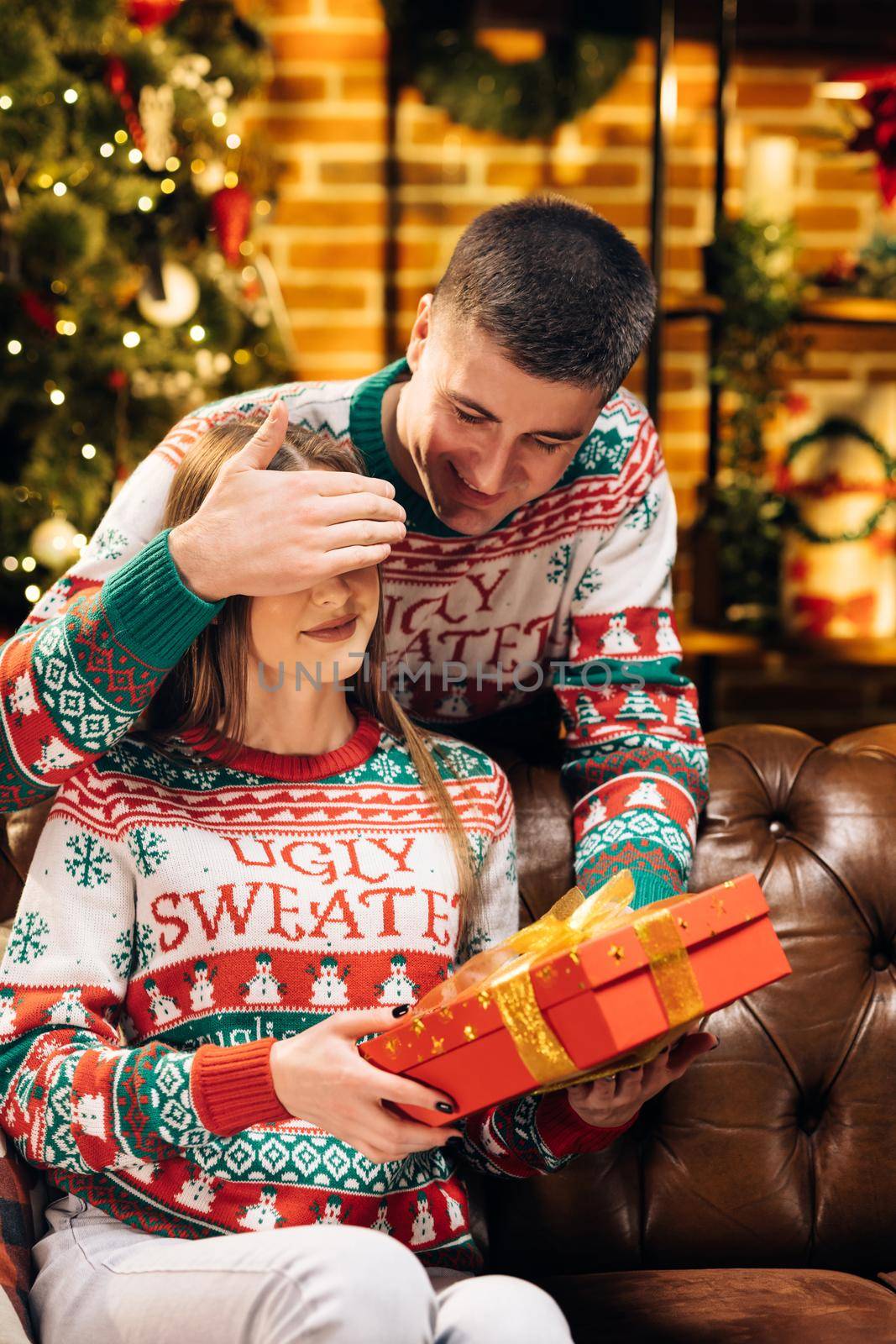 Happy surprised woman receiving christmas gift from loving man at home on Christmas Eve near decorated xmas tree. Caucasian male giving present to beloved female woman on holidays. Family celebration.