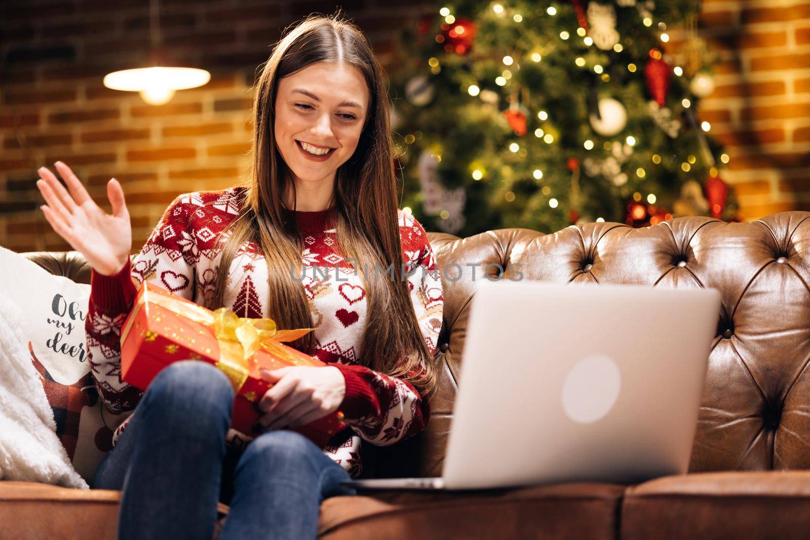 Excited happy caucasian girl wears santa hat holds xmas gift box, opens Christmas present on computer video call virtual meeting chat with family, social distance friend sits on sofa at home.