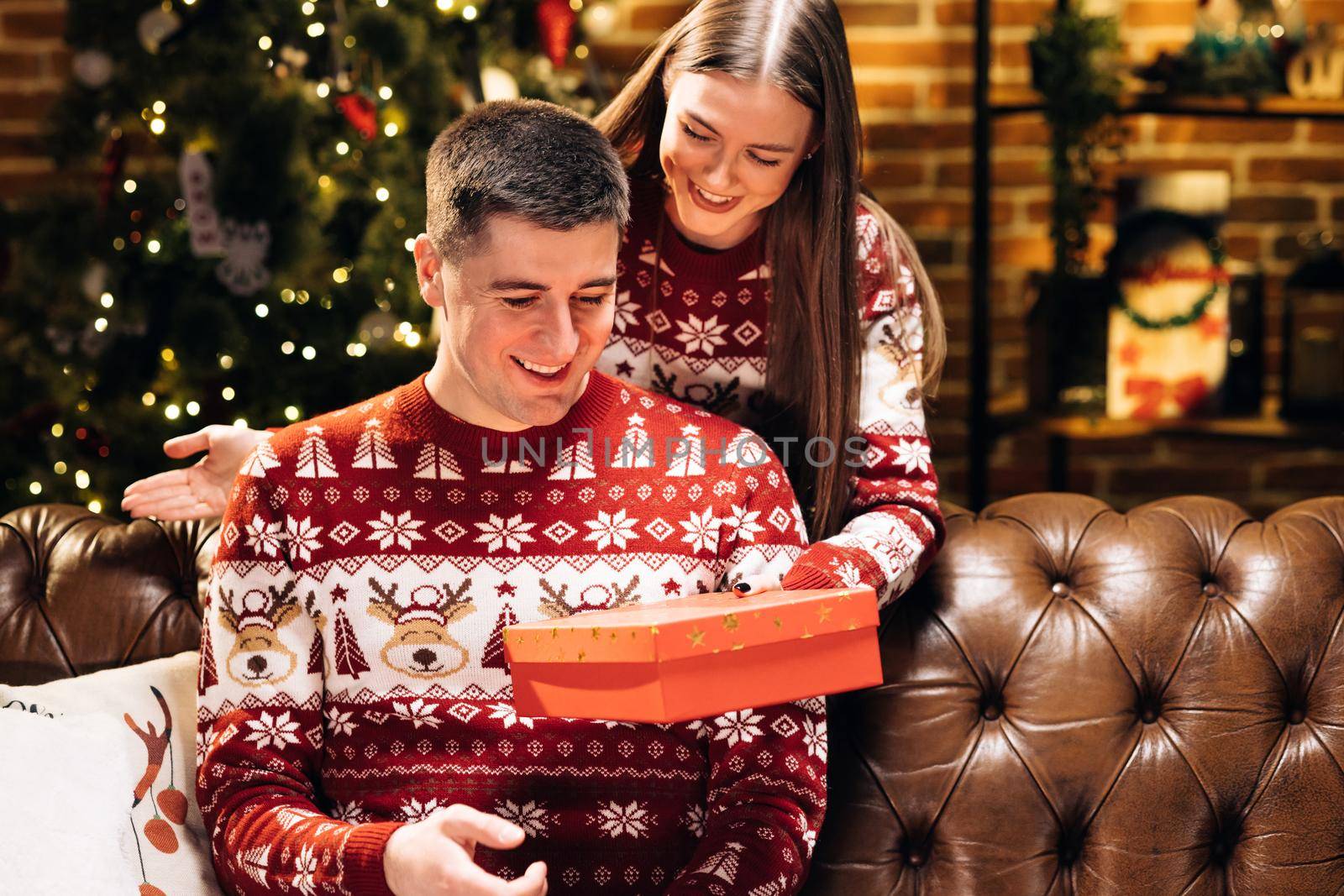 Young 30s woman covering eyes of smiling curious husband, giving wrapped box with Christmas gift near decorated festive tree, New Year winter holidays family celebration. Holiday miracle by uflypro