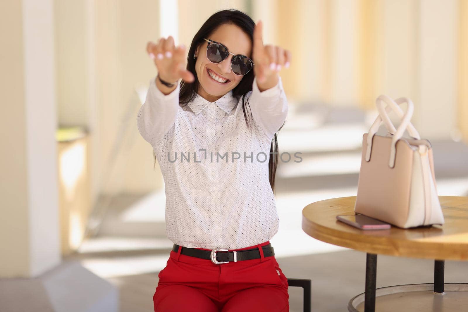 Portrait of cheerful happy young woman in stylish outfit show some symbols with hands. Attractive lady sit outdoors, gesture on camera. Trendy, joy concept