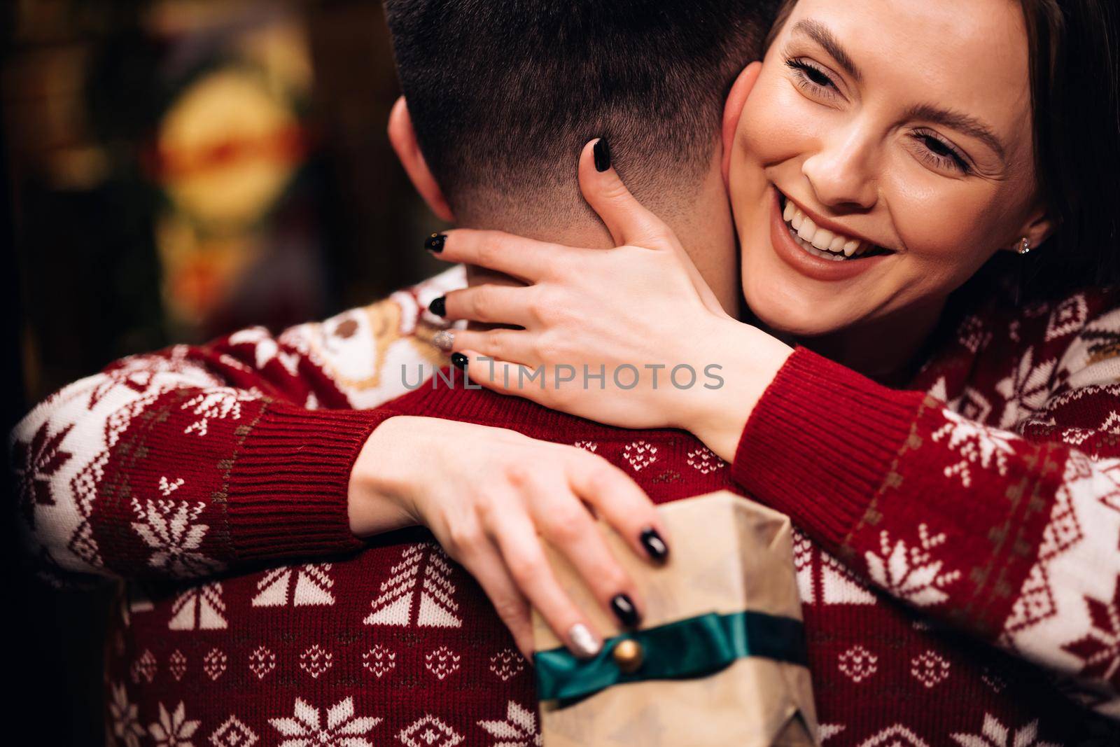 Young happy woman embracing her boyfriend and hugging him near decorated glowing fir tree. Romantic winter story. Happy wife feels grateful hugs husband express gratitude for New Year gift.