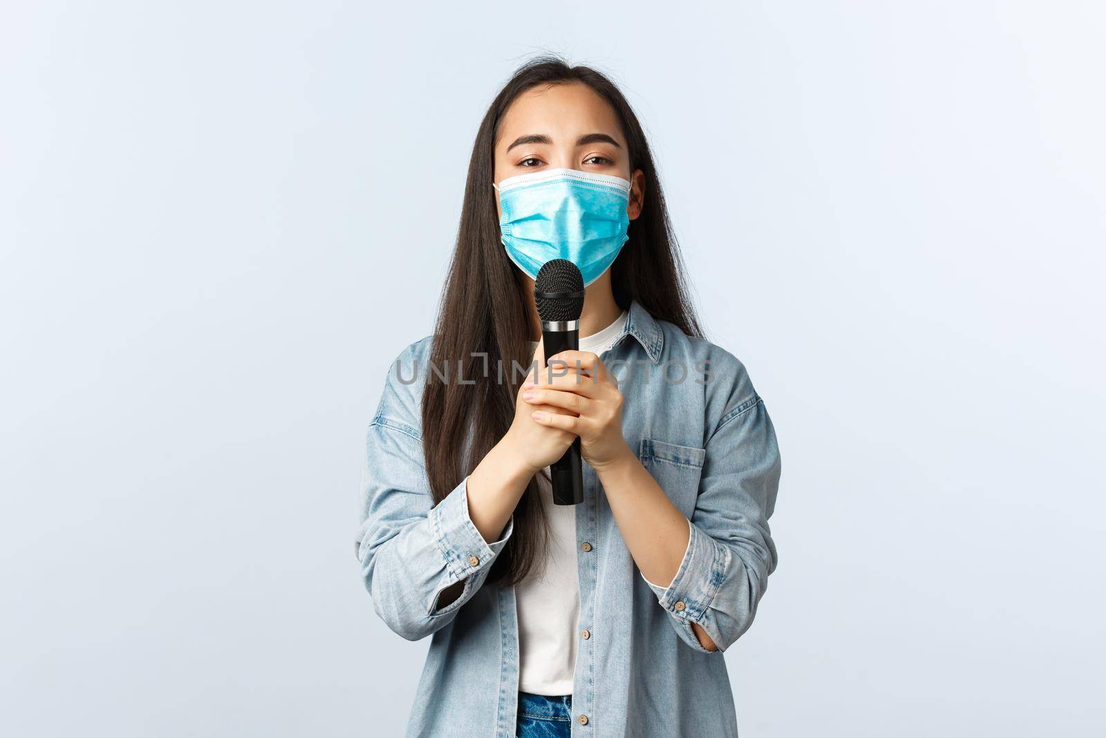 Social distancing lifestyle, covid-19 pandemic and self-isolation leisure concept. Carefree passionate asian woman in medical mask singing with microphone, close eyes delighted.