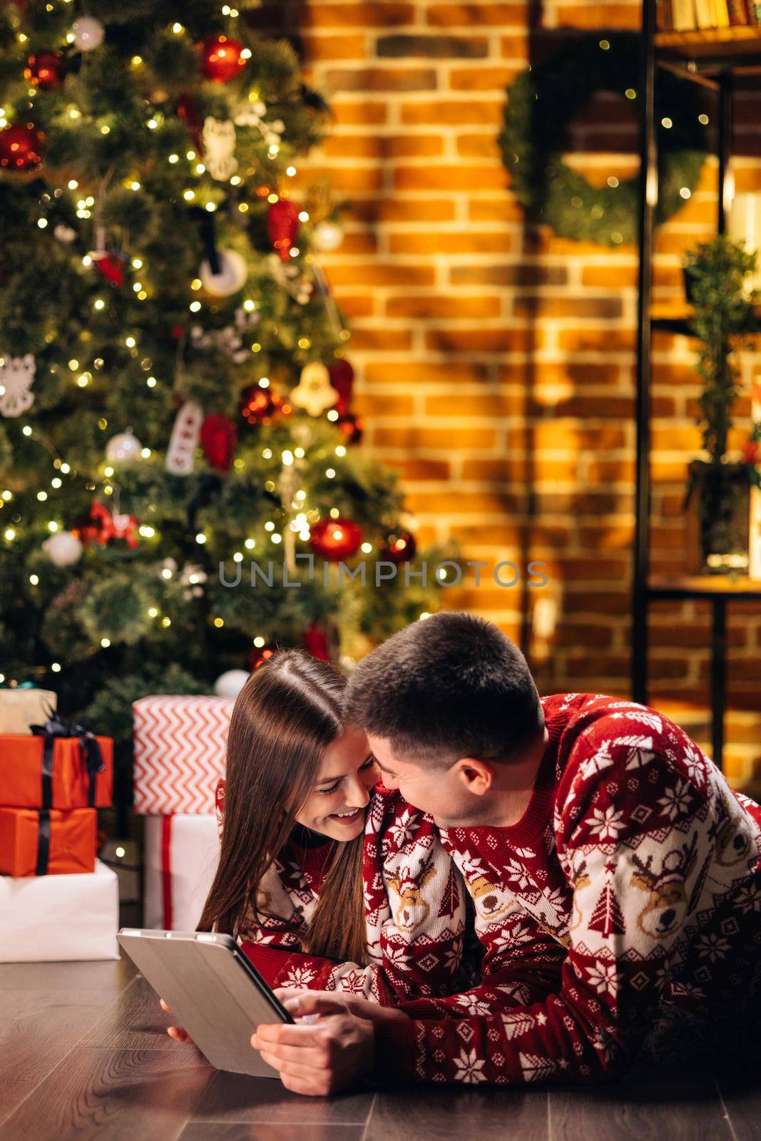 Cheerful caucasian couple man and woman resting at home near glowing new year tree and typing on tablet looking for xmas presents online on holiday sales. Buy online, sale, modern tech overuse concept