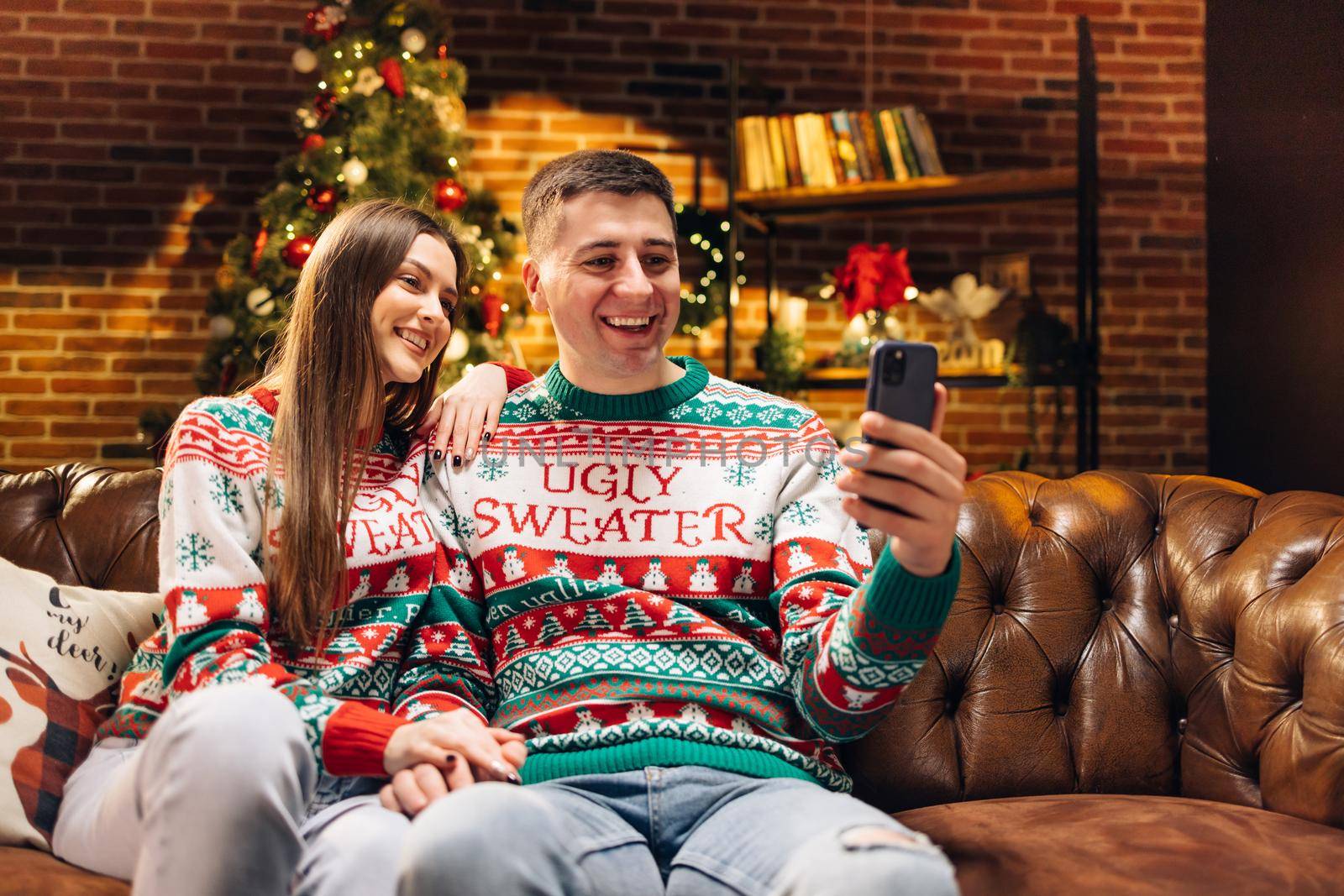 Attractive Man and Woman Talking by Video Call Sending Positive Emotions Looking at Camera. Happy Couple Sitting at Home while Recording Christmas Greetings. Happy New Year. Merry Christmas