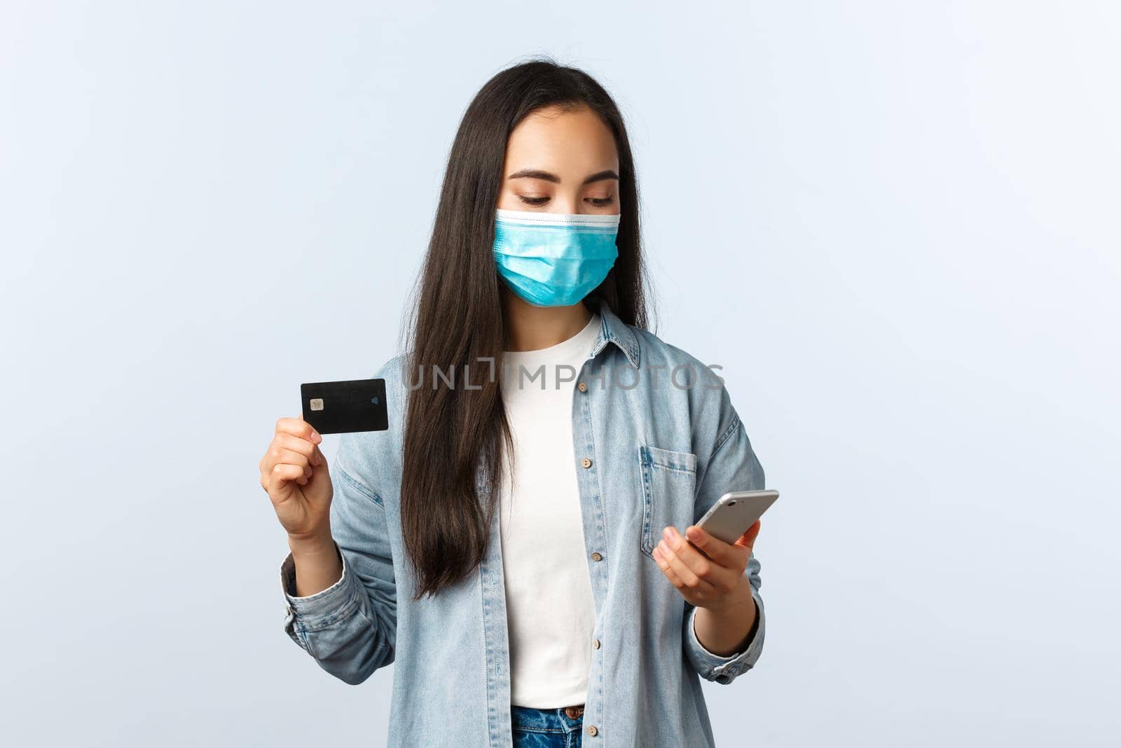 Social distancing lifestyle, covid-19 pandemic and contactless shopping concept. Cute teenage asian girl in medical mask, show credit card and looking at mobile phone while making order.