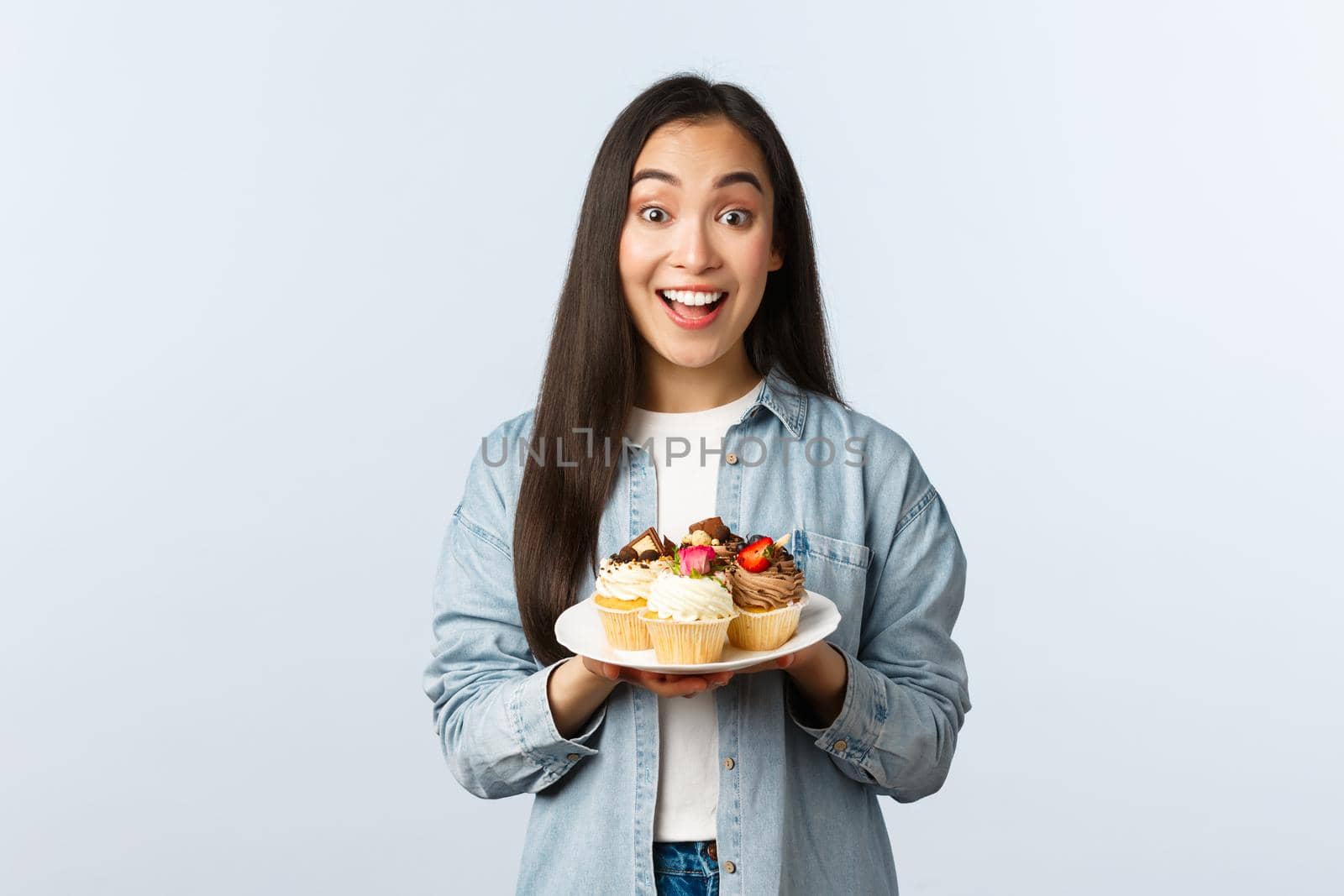 Social distancing lifestyle, covid-19 pandemic, celebrating holidays during coronavirus concept. Excited asian girl holding plate with tasty cupcakes, smiling broadly, invite visit cafe with muffins.