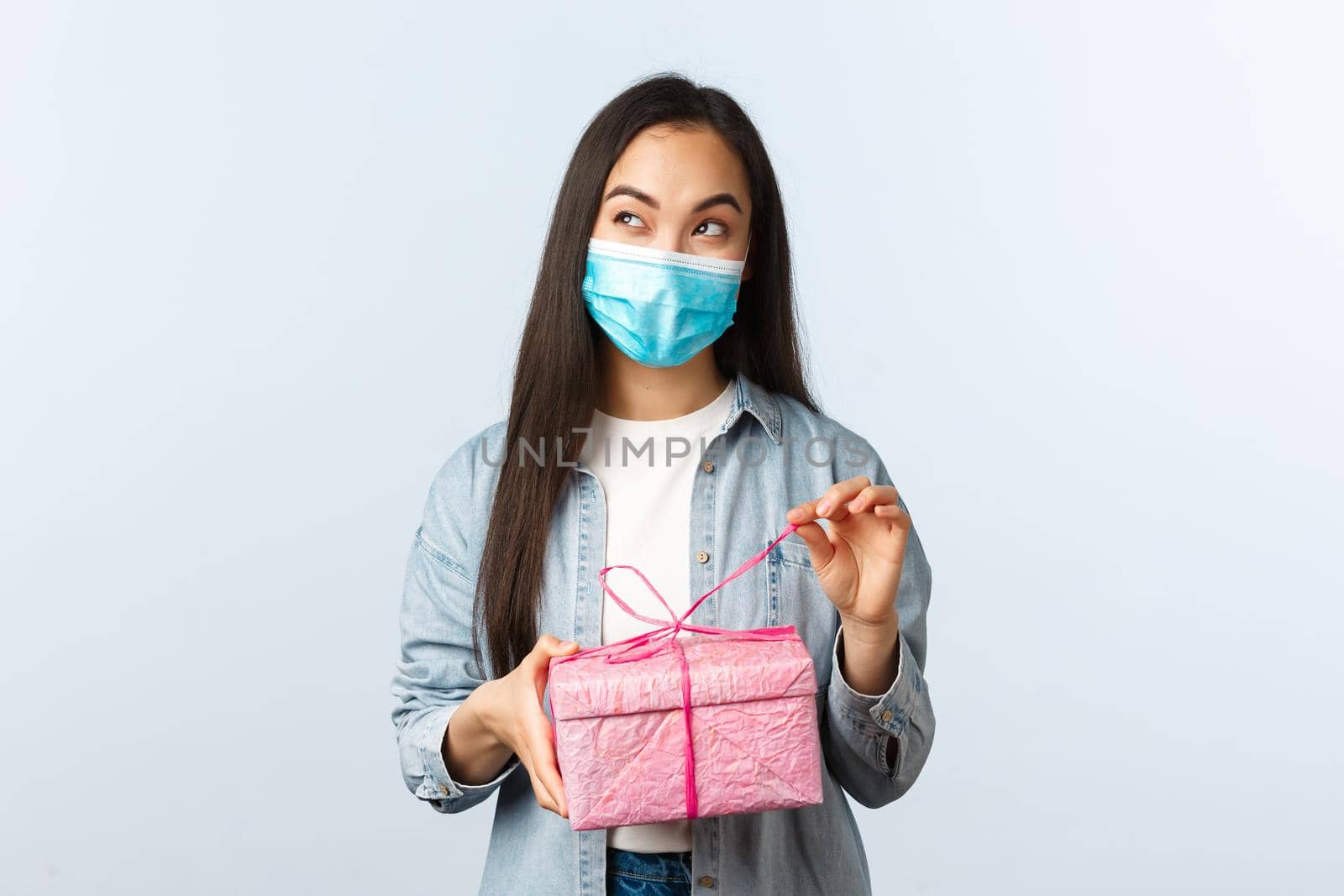 Social distancing lifestyle, covid-19 pandemic, celebrating holidays during coronavirus concept. Intrigued happy asian woman in medical mask unwrap gift box thinking what inside by Benzoix