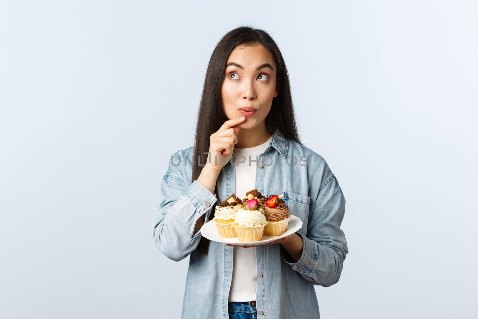 Social distancing lifestyle, covid-19 pandemic, celebrating holidays during coronavirus concept. Dreamy asian girl thinking eat cupcake or not, look away thoughtful, holding muffin plate.