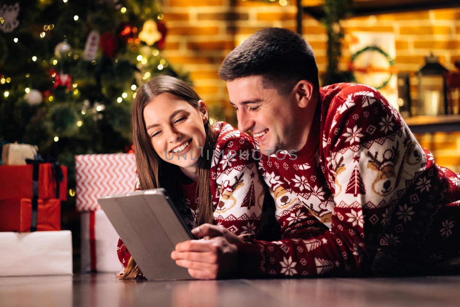 Happy caucasian couple lying home near Christmas tree background. Beautiful woman with long hair holds digital tablet and emotionally tells funny stories by uflypro