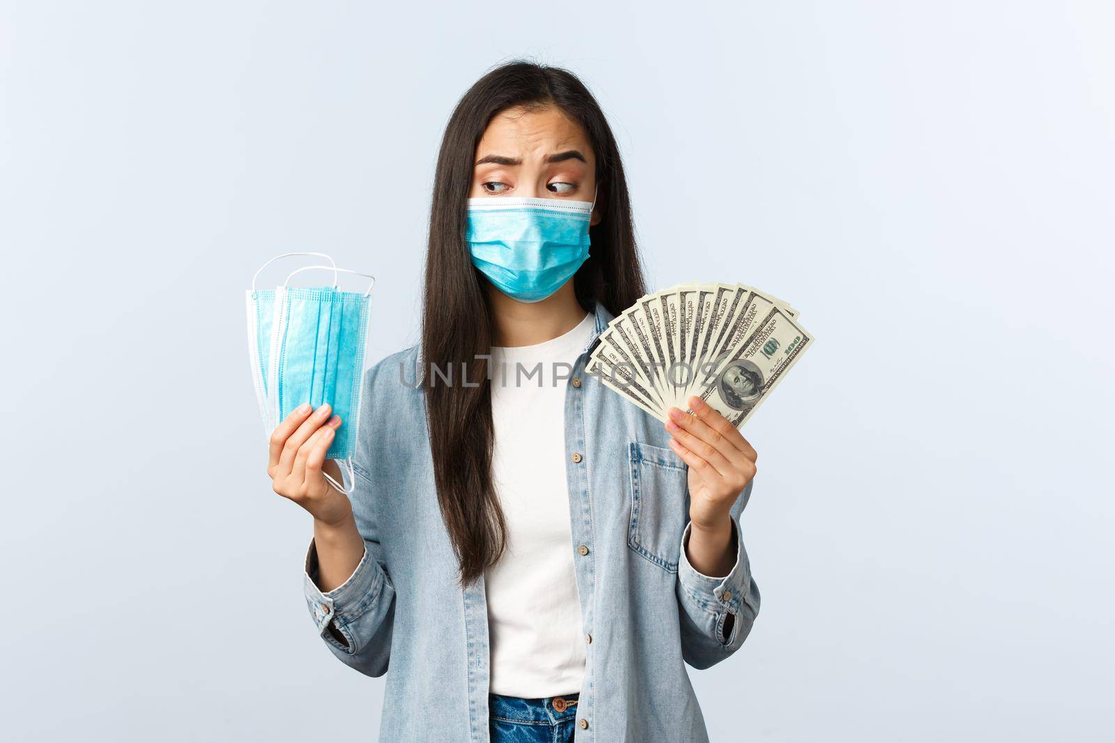 covid-19 pandemic, coronavirus expences and finance concept. Thoughtful and puzzled asian female looking at medical mask and showing money, losing money during coronavirus.