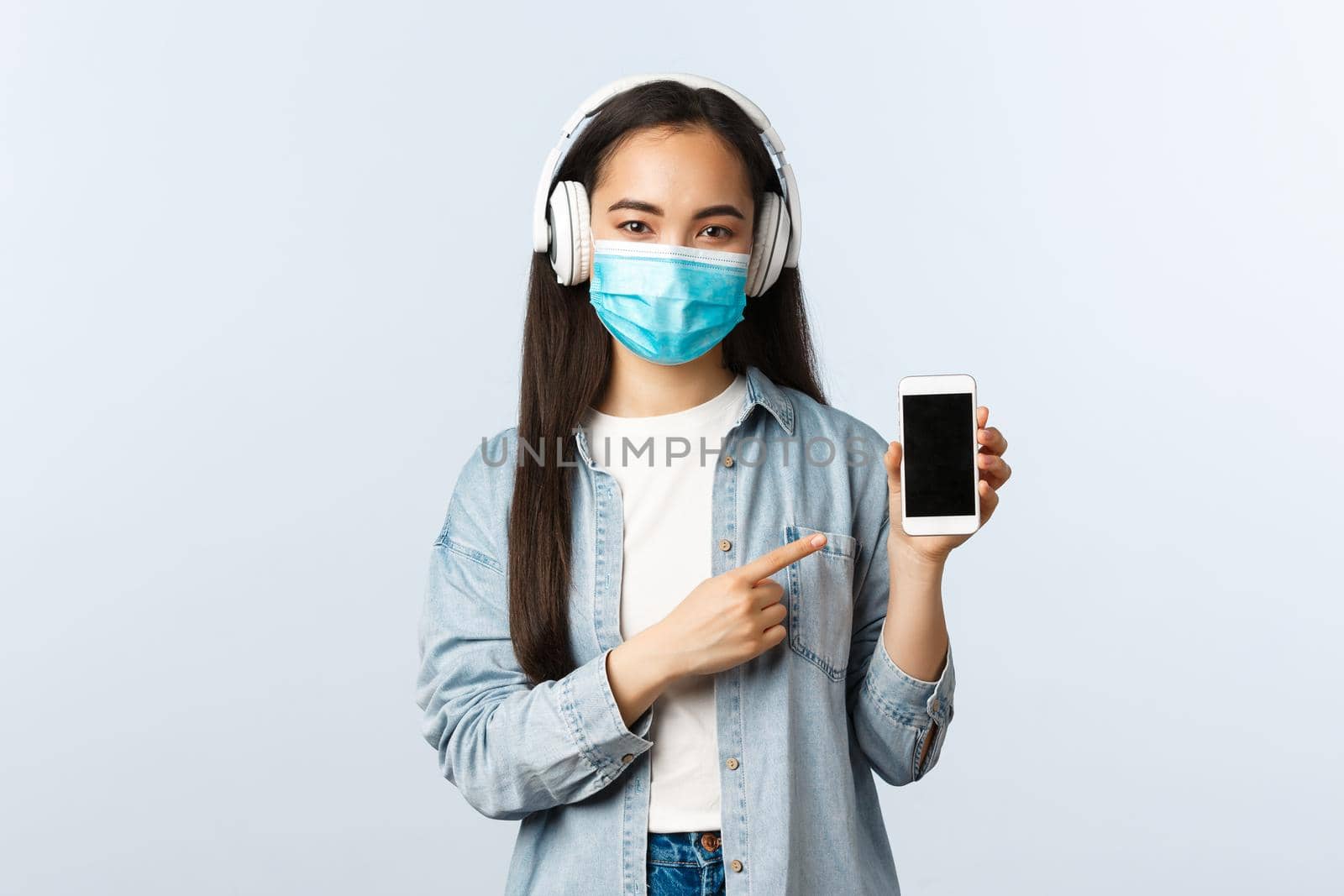 Social distancing lifestyle, covid-19 pandemic and self-isolation leisure concept. Smiling stylish teenage asian girl listening music in wireless headphones, pointing mobile phone screen.