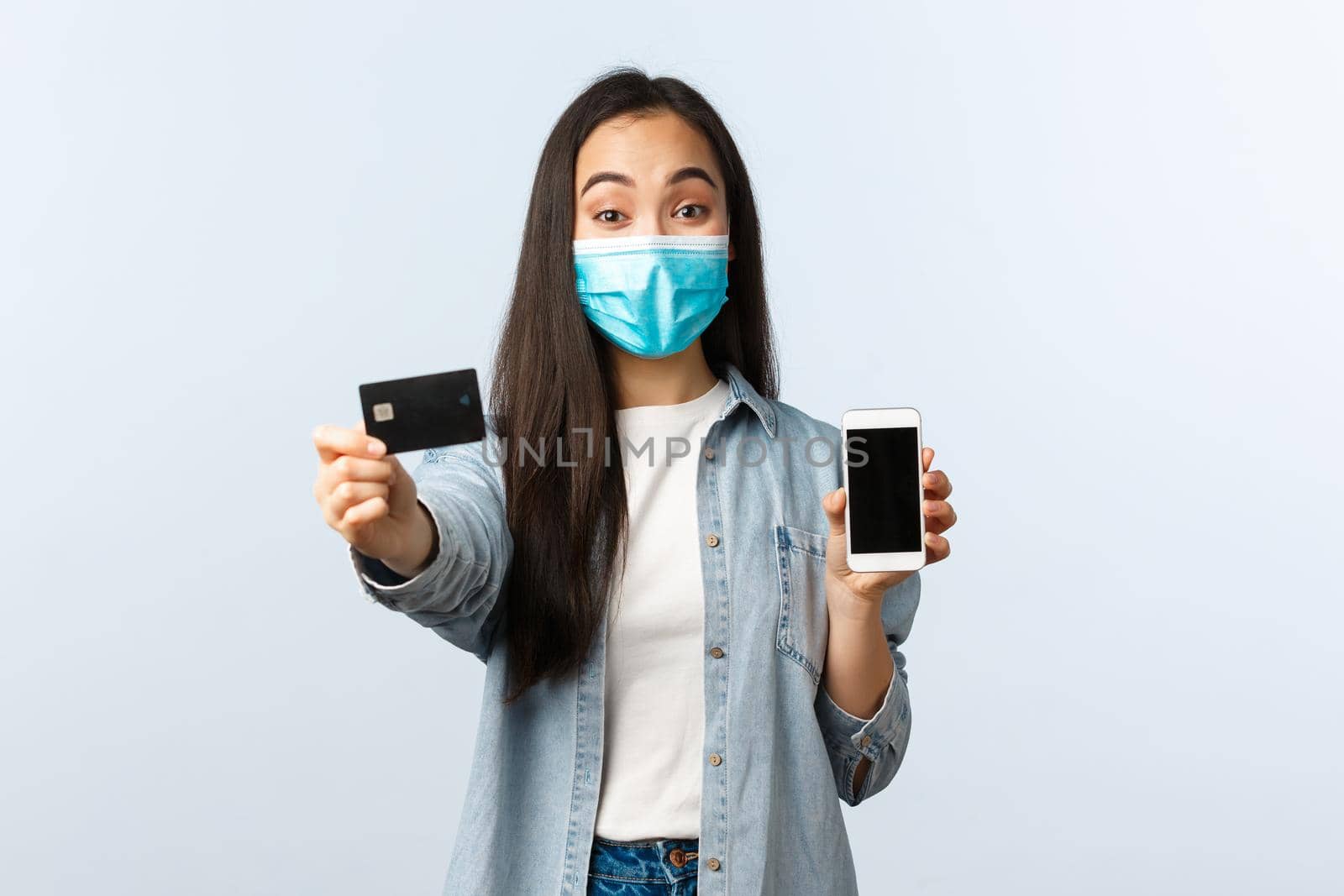 Social distancing lifestyle, covid-19 pandemic and contactless shopping concept. Cheerful pretty asian girl in medical mask advice using credit card for paying online for orders by Benzoix