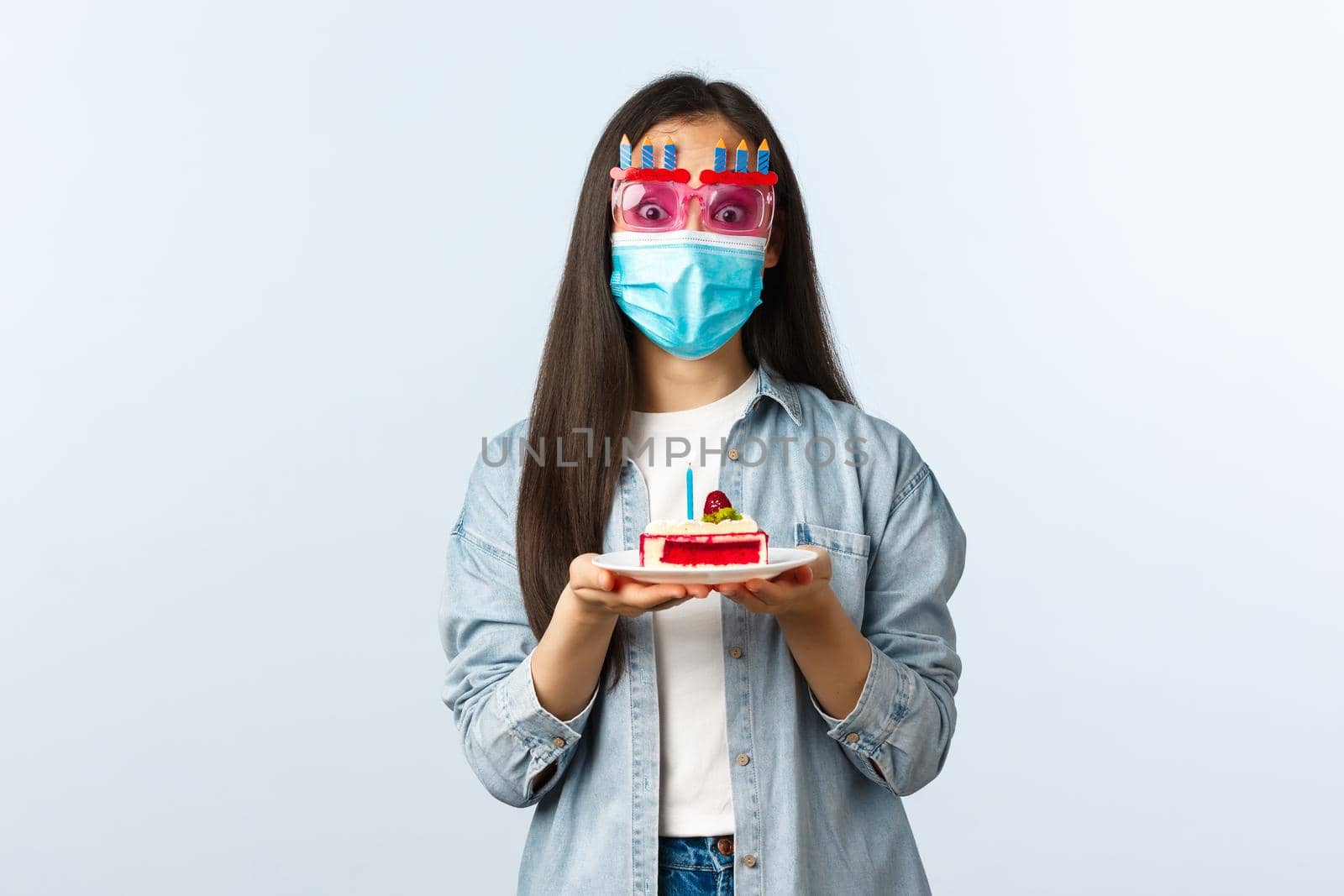 Social distancing lifestyle, covid-19 pandemic, celebrating holidays during coronavirus concept. Cheerful birthday girl in medical mask and party glasses, holding b-day cake, enjoying celebration by Benzoix
