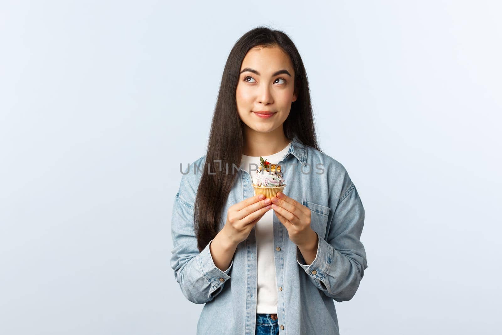Social distancing lifestyle, covid-19 pandemic, celebrating holidays during coronavirus concept. Thoughtful cute asian girl deciding eat cupcake or note, look up thinking, holding delicious dessert by Benzoix