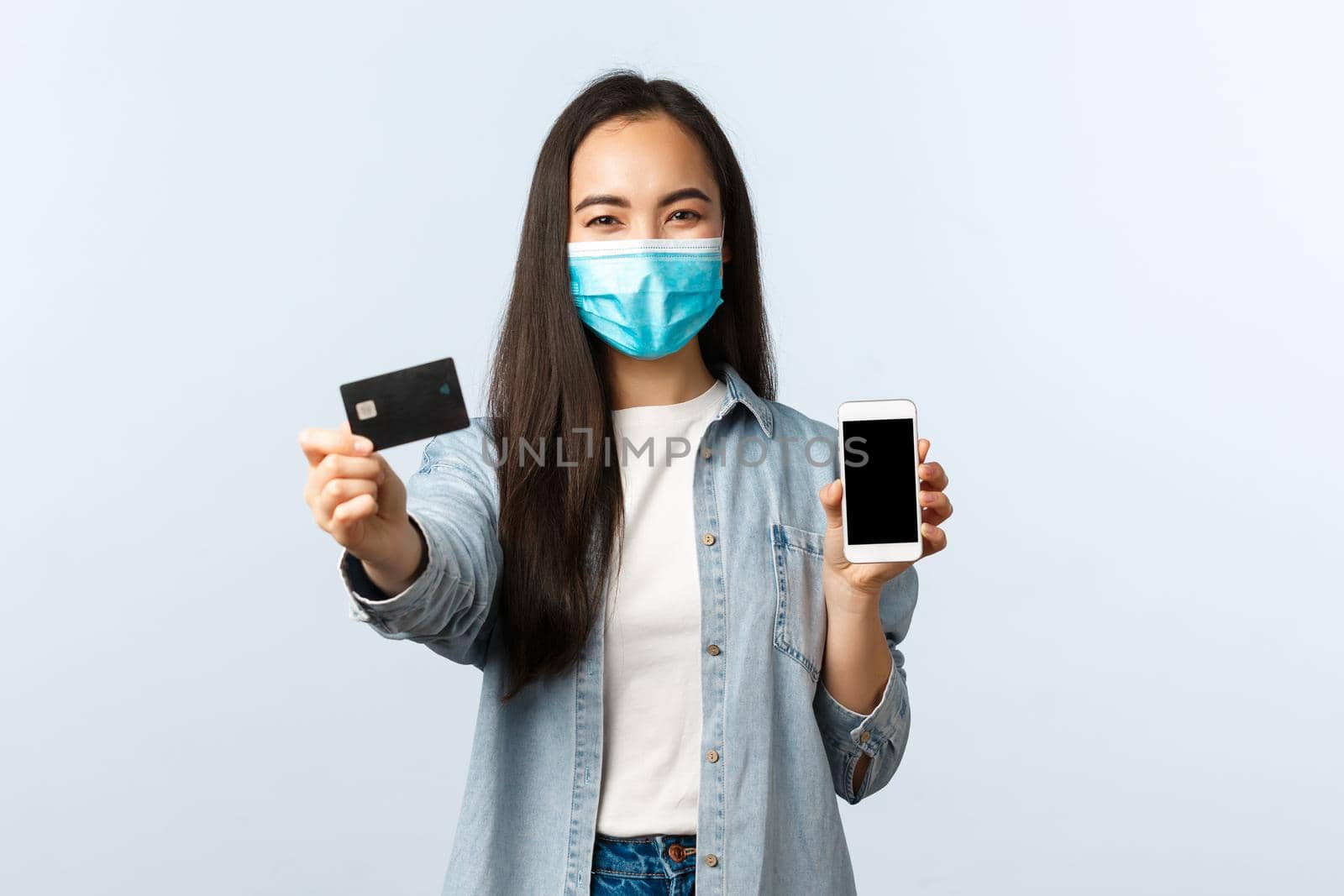 Social distancing lifestyle, covid-19 pandemic and contactless shopping concept. Cheerful friend asian girl in medical mask showing credit card and mobile screen, advice order online and pay by Benzoix