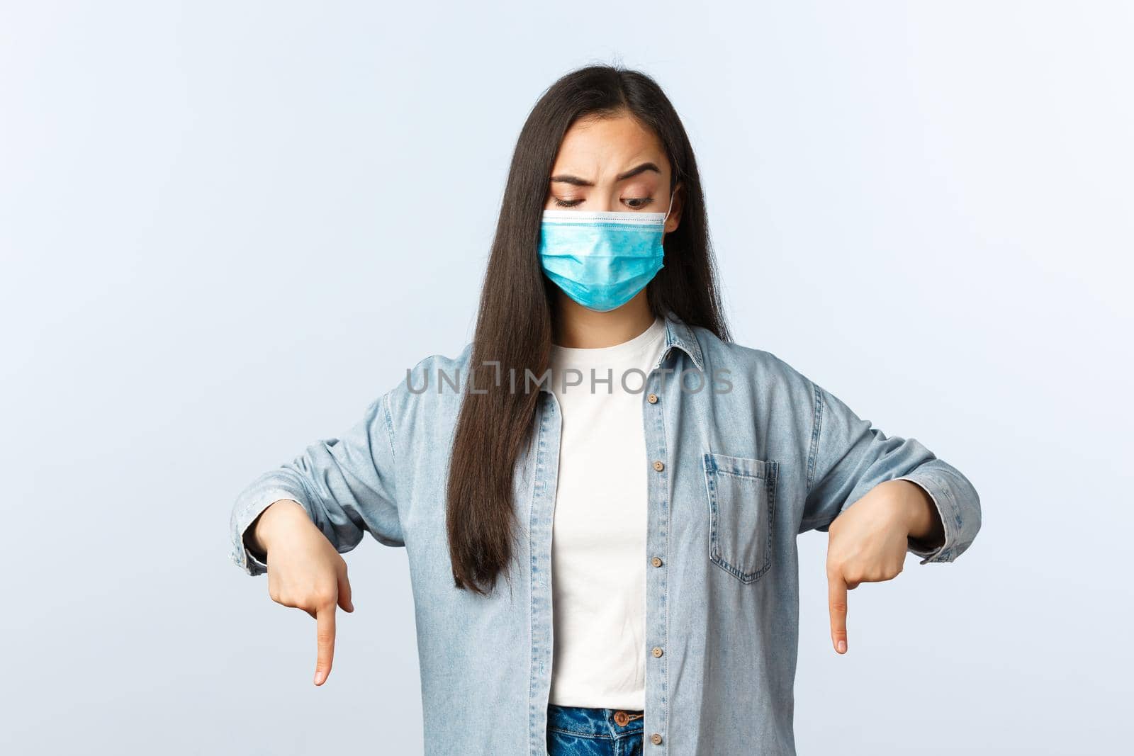 Social distancing lifestyle, covid-19 pandemic everyday life and leisure concept. Skeptical and confused asian woman in medical mask, casual clothes, looking and pointing down by Benzoix